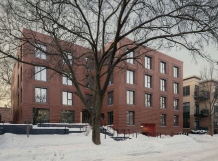A modern, four-story brick apartment building with large windows, set behind a leafless tree and surrounded by snow on the ground and sidewalks.