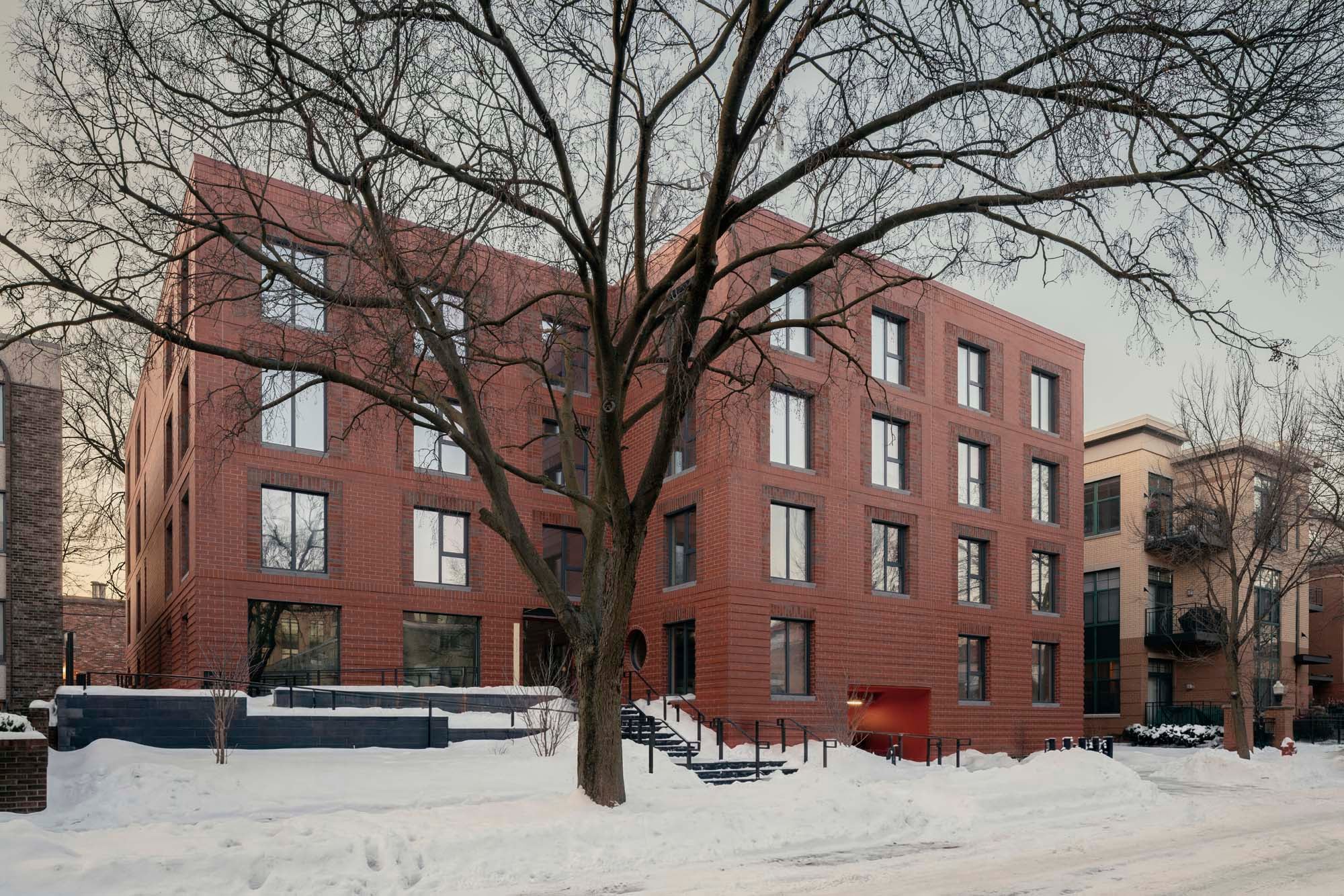 A modern, four-story brick apartment building with large windows, set behind a leafless tree and surrounded by snow on the ground and sidewalks.