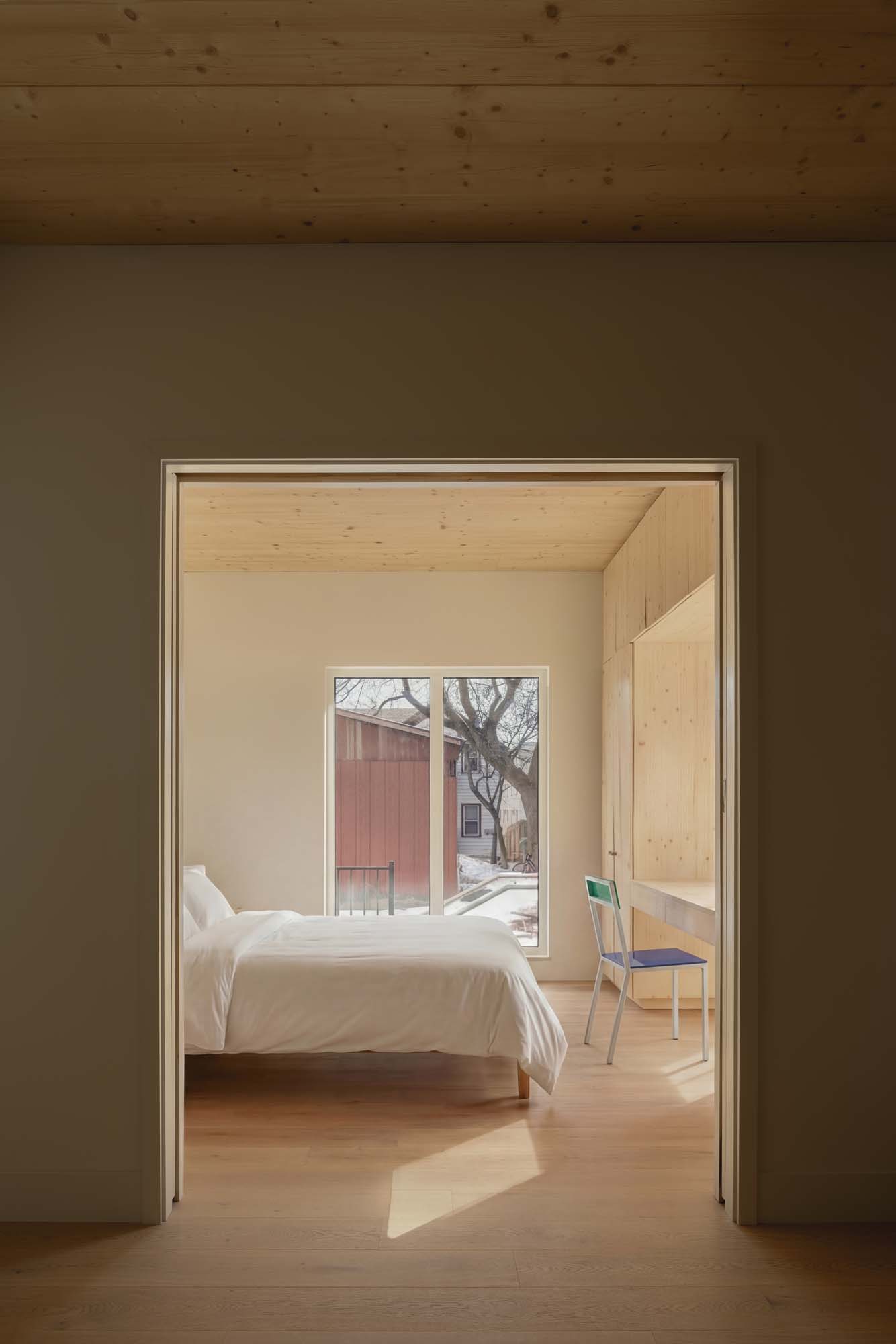 A minimalist bedroom with a wooden ceiling and floor, a white bed, large window, built-in desk, and a colorful chair. Sunlight enters through the window.