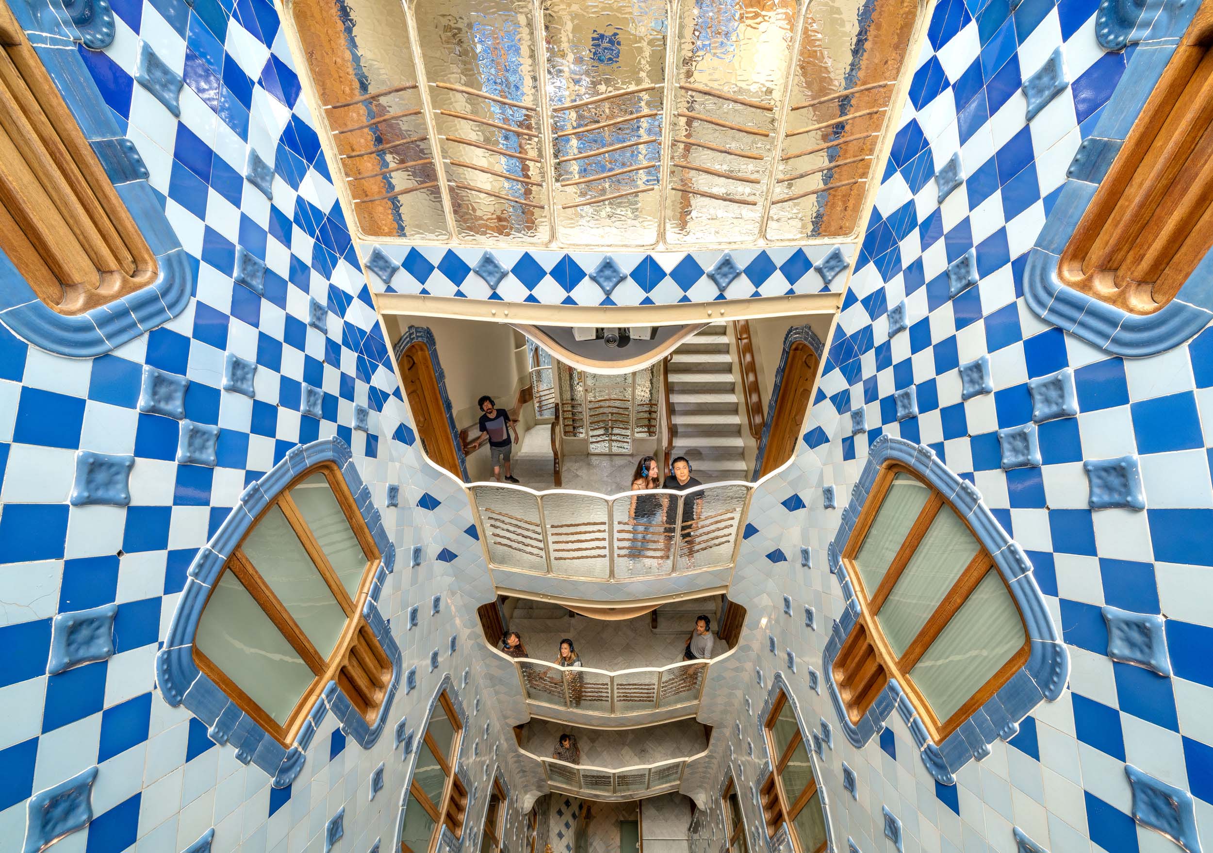 People look out from balconies inside a multistory building with blue and white tiled walls, arched windows, and decorative railings inspired by Gaudi, viewed from below.