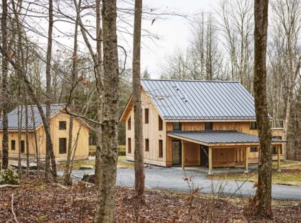 Two modern wooden houses with metal roofs are situated in a leafless forest setting, connected by a gravel driveway on an overcast day.