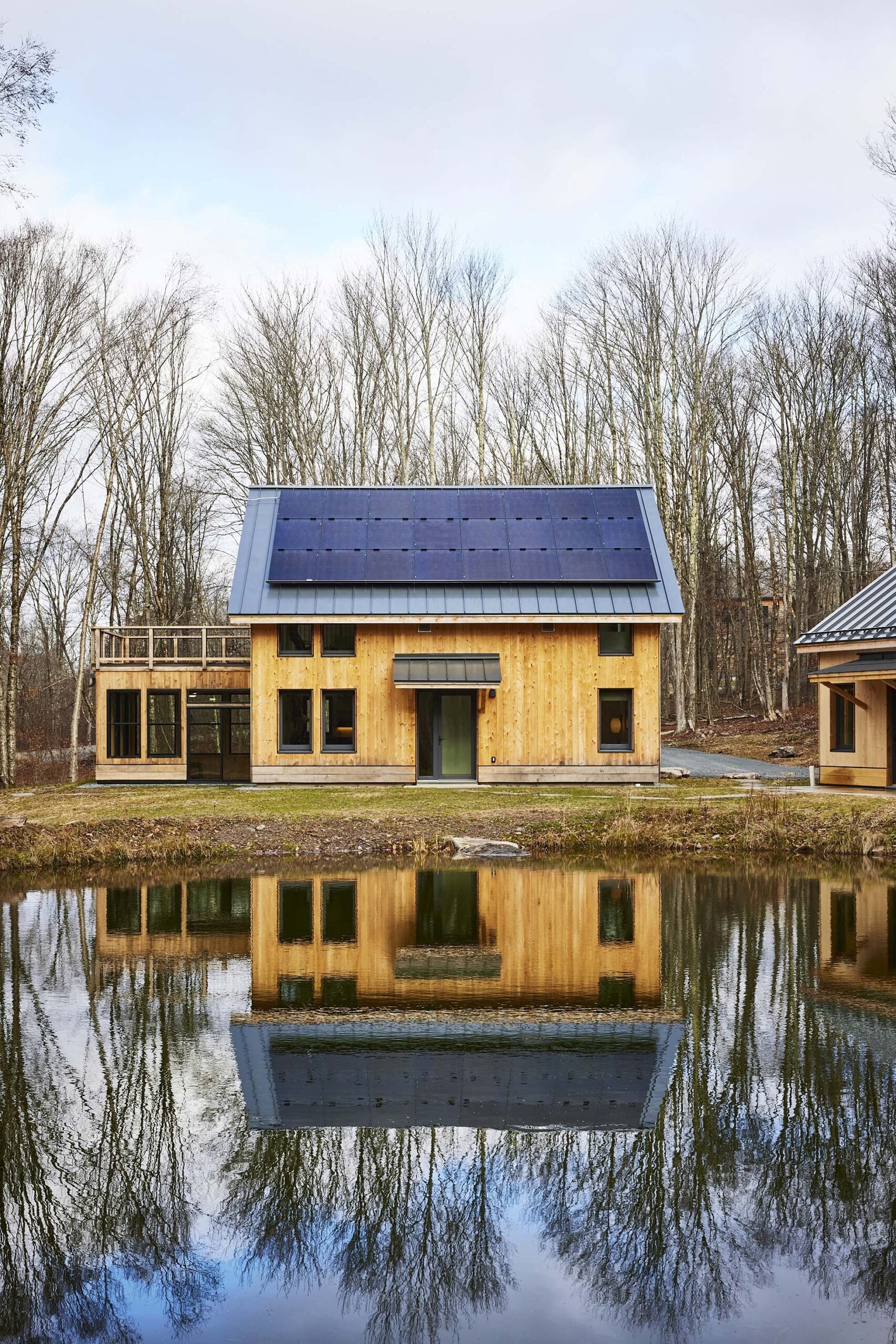 A modern wooden house with solar panels on the roof is reflected in a small pond, surrounded by leafless trees and cloudy sky.
