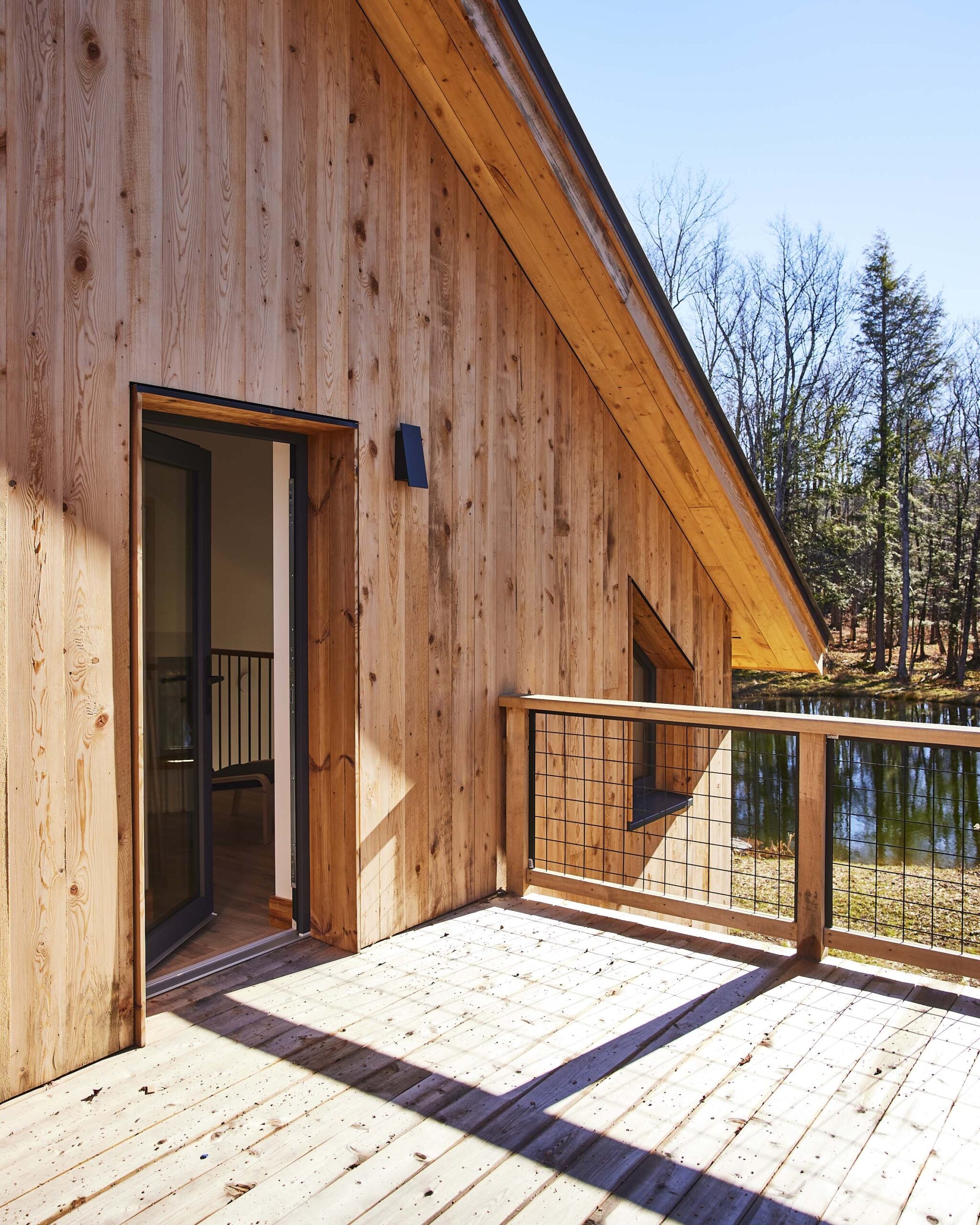 A wooden balcony with a metal railing extends from a house, overlooking a pond and trees on a clear, sunny day.