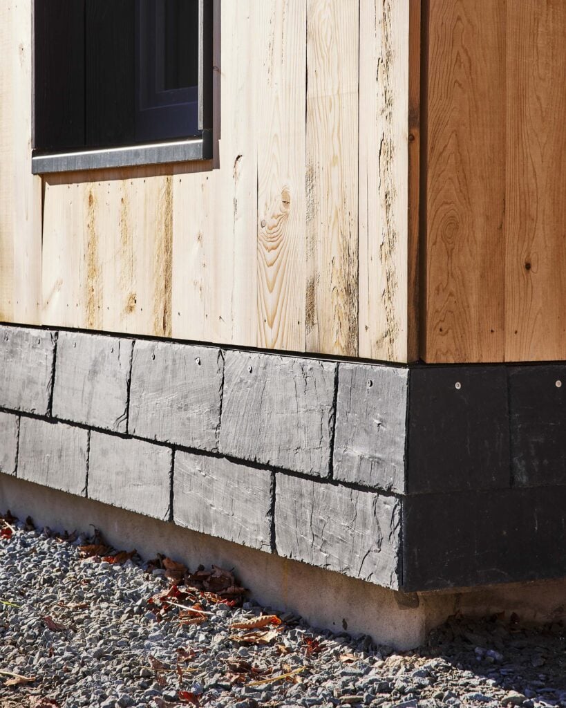 Close-up of a building’s exterior corner showing vertical wooden siding above a row of gray stone or concrete blocks and a gravel foundation.