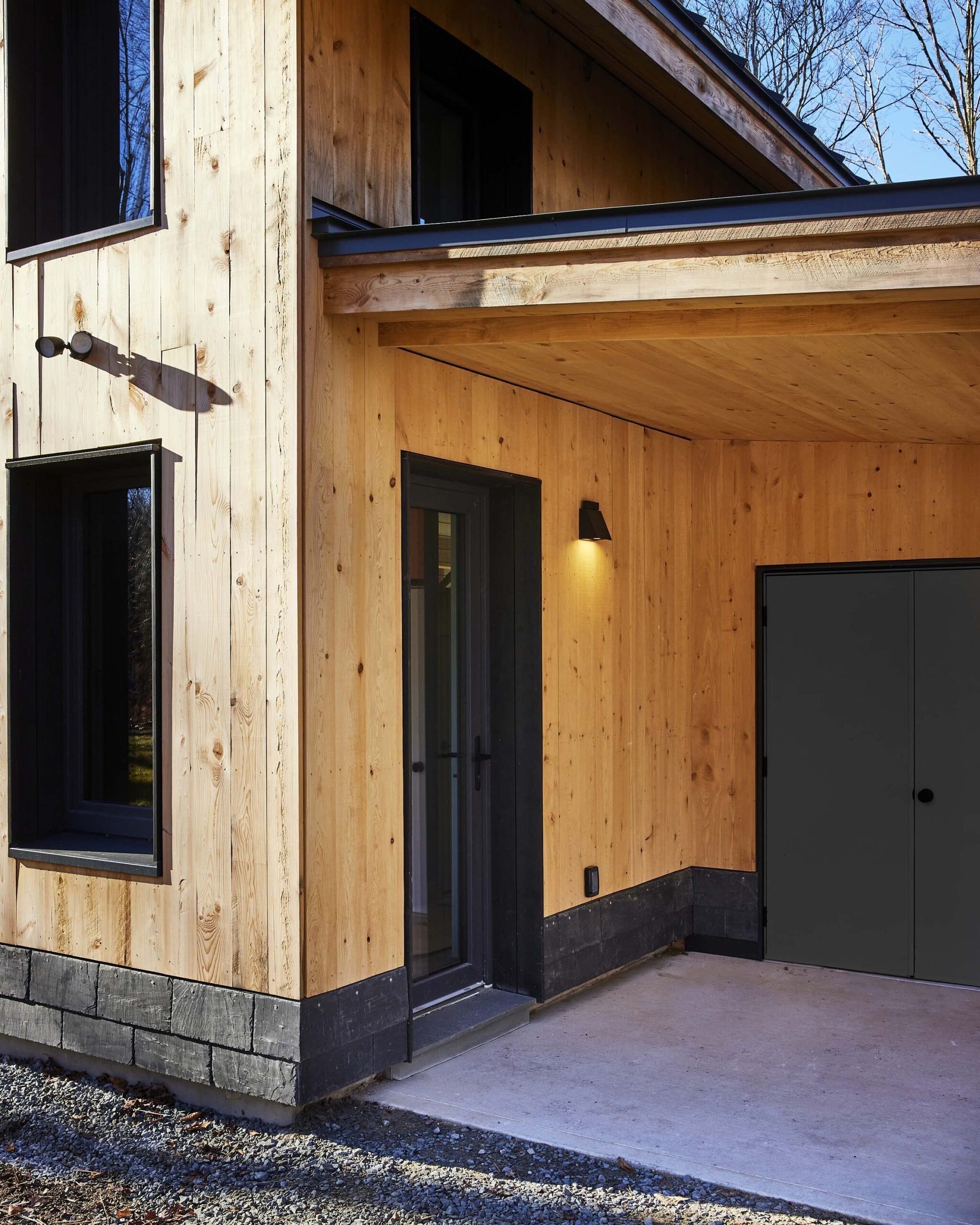 Modern house exterior with light wood siding, black trim, a covered entryway, black door, and outdoor wall lights.