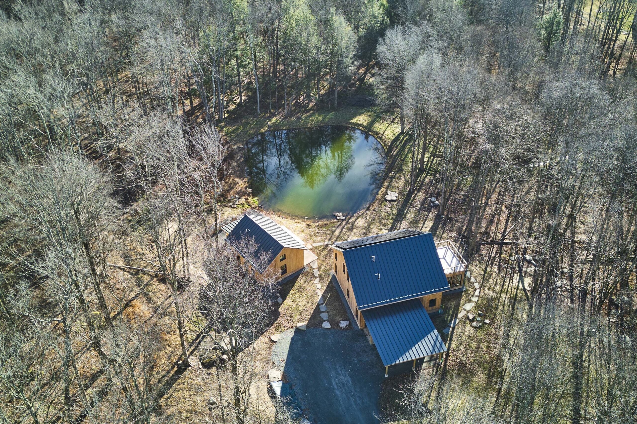 Aerial view of two wooden cabins with blue roofs near a small pond surrounded by leafless trees in a forested area.