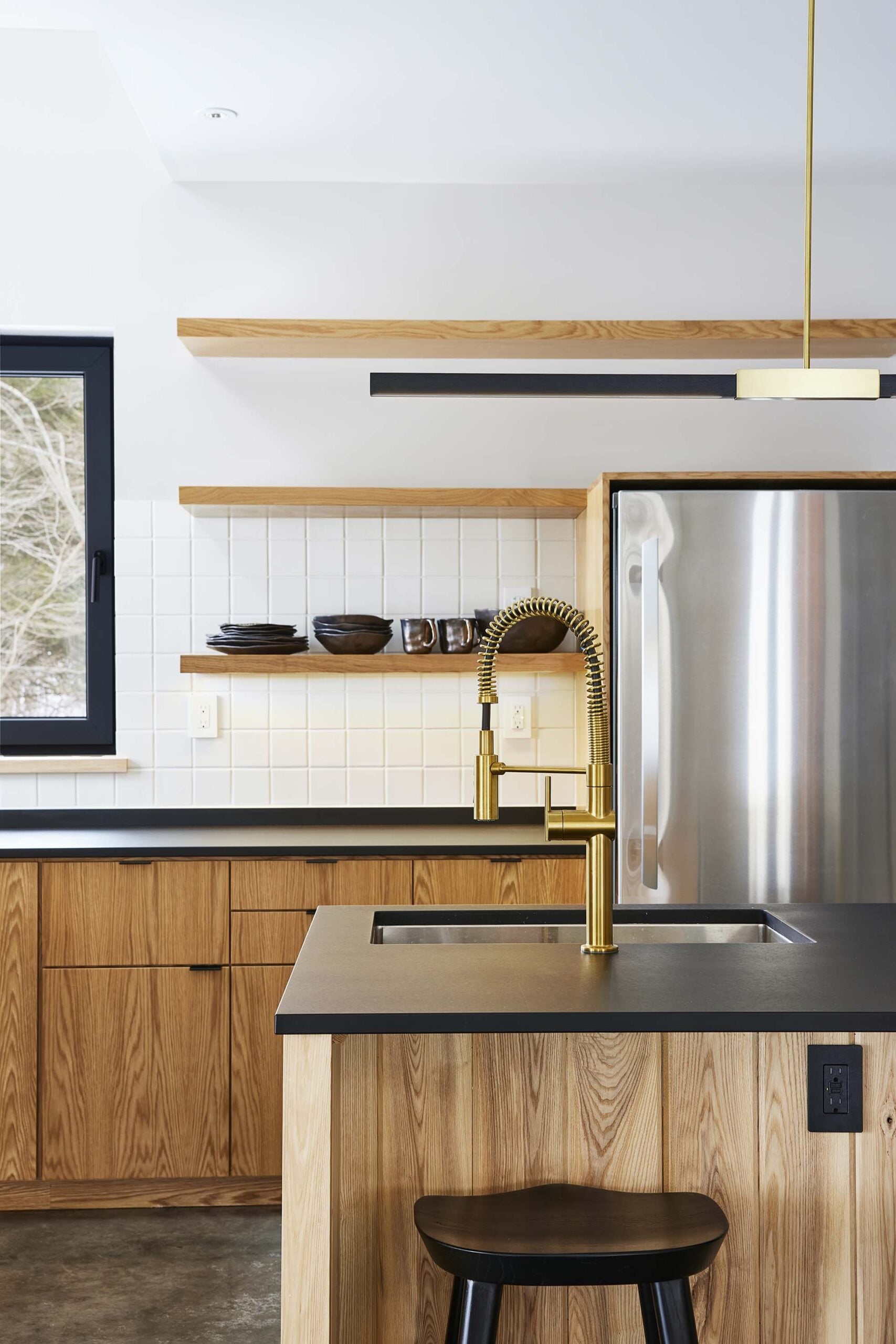 Modern kitchen with wood cabinets, black countertops, open shelves with dishes, a stainless steel fridge, and a gold pull-down faucet at a central island sink.