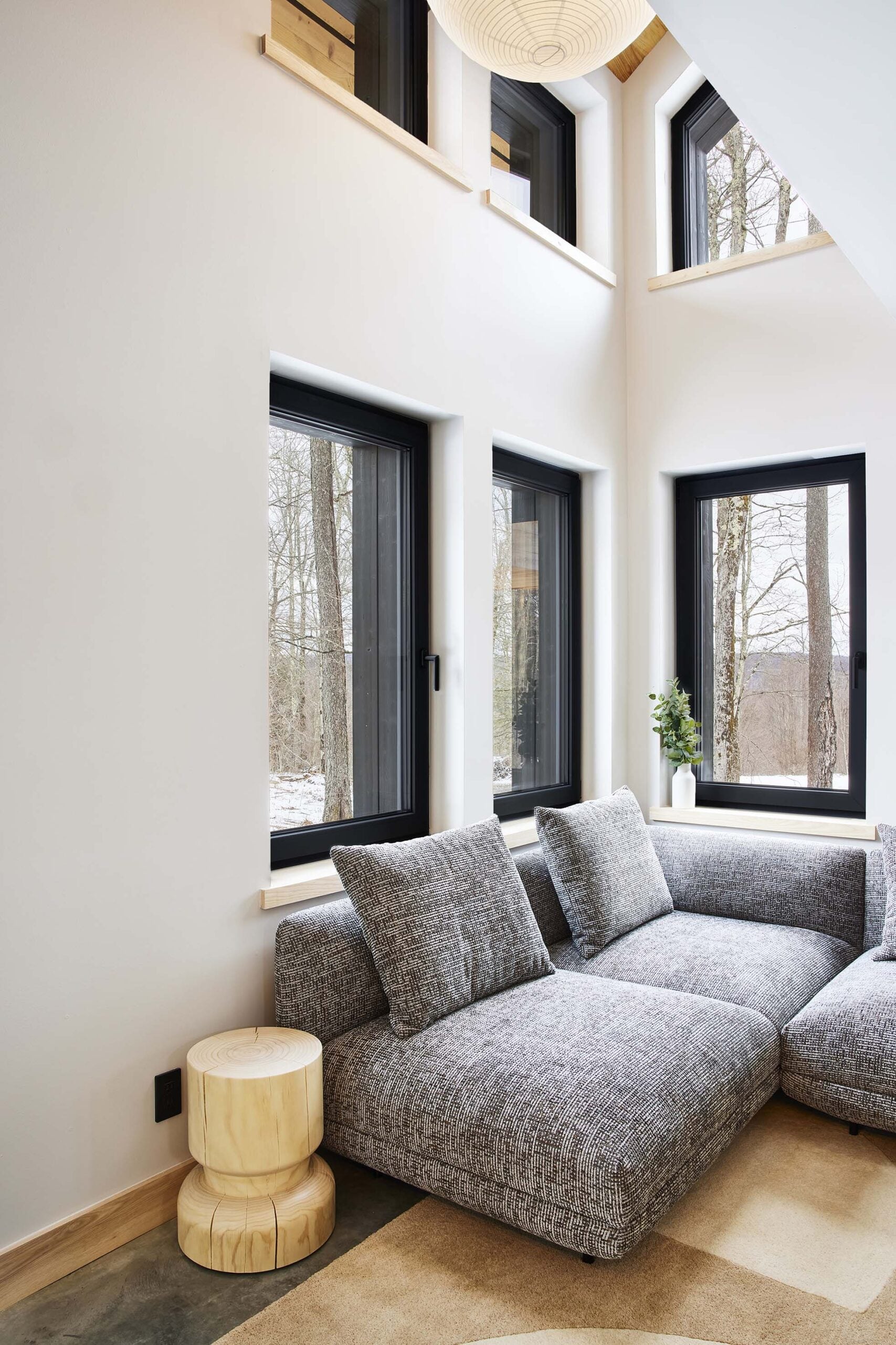 Modern living room with large black-framed windows, a gray sectional sofa, light wood side tables, and views of trees outside.