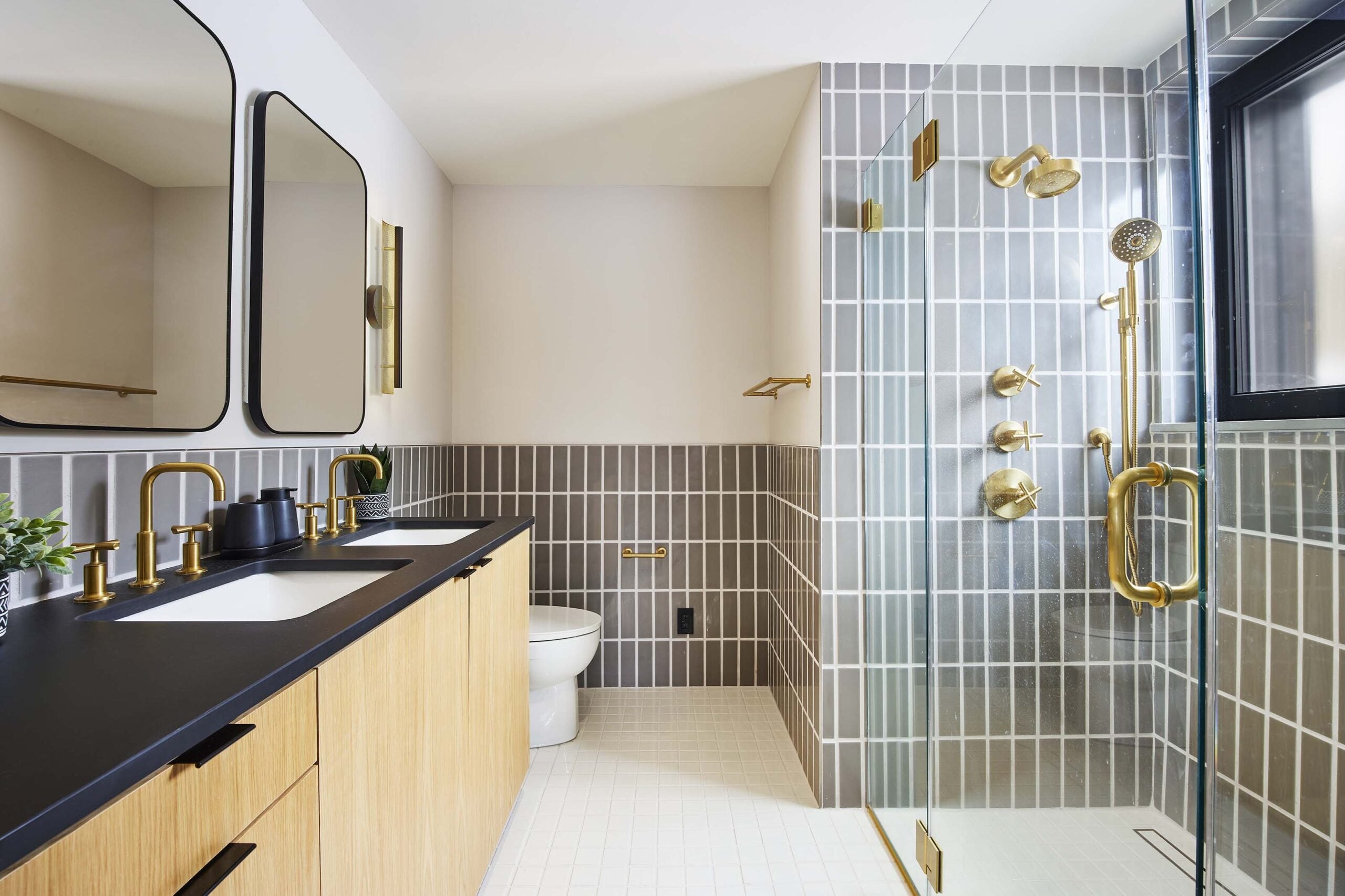 Modern bathroom with dual sinks, black countertop, light wood cabinets, gray tiled walls, glass shower enclosure, brass fixtures, and a toilet.