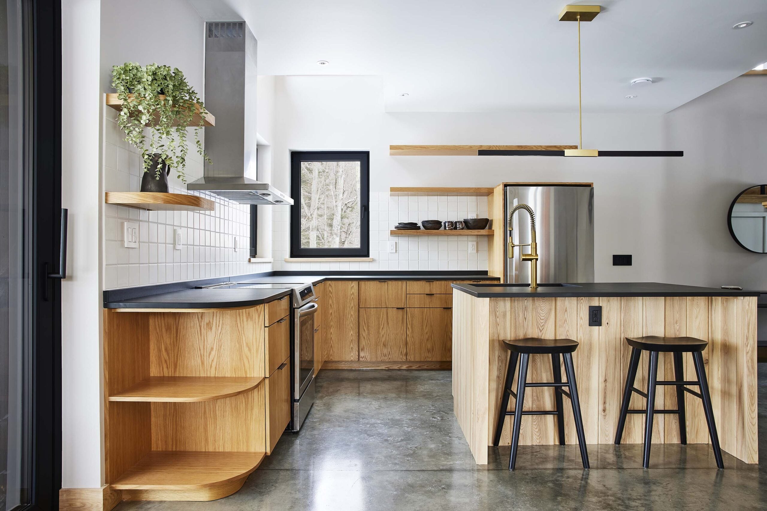Modern kitchen with wood cabinetry, black countertops, open shelves, stainless steel appliances, a potted plant on a shelf, and two black barstools at the island.