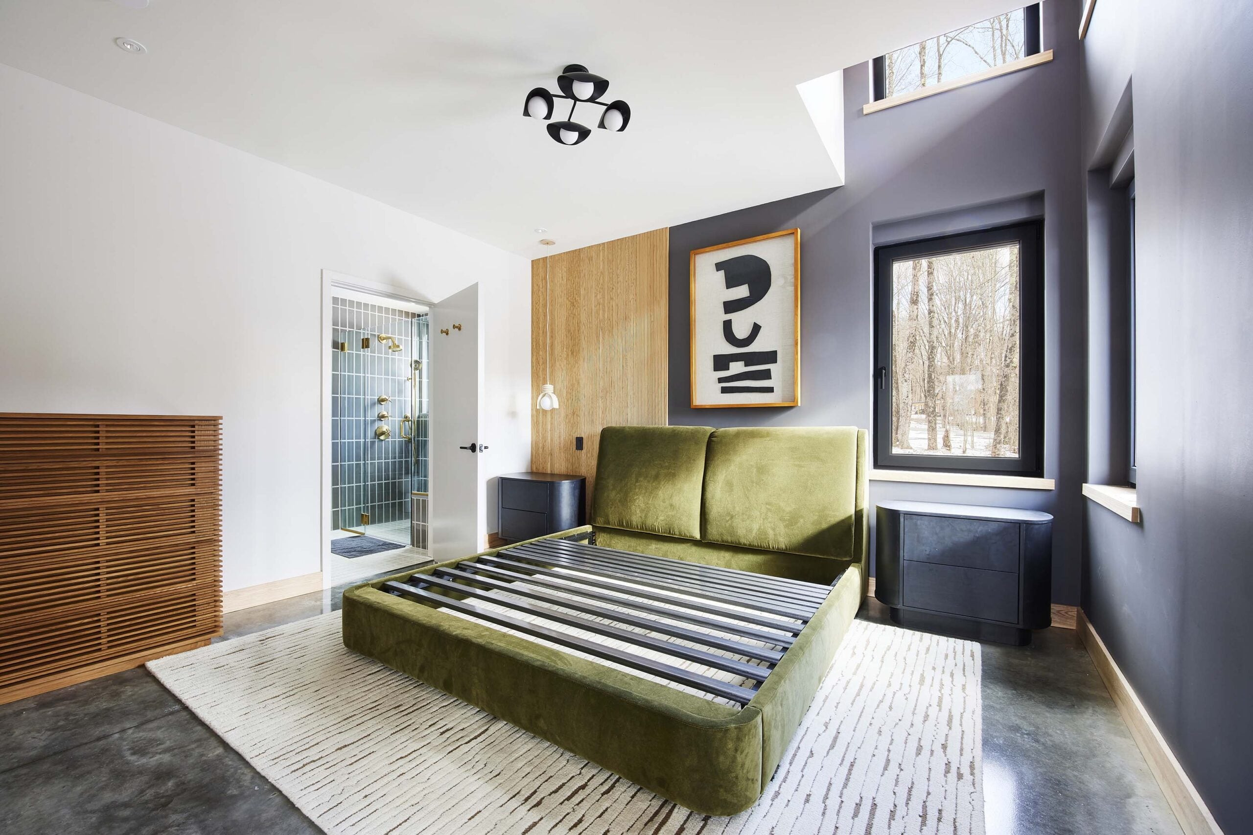 Modern bedroom with a green velvet bed frame without a mattress, abstract wall art, large windows, and an adjacent bathroom with glass shower doors.