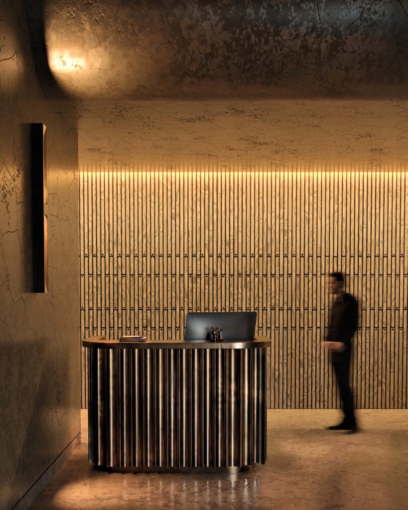 A reception desk with a computer and two small decor items sits in front of a textured wall; a blurred person walks by on the right side. Warm lighting highlights the scene, capturing urban elegance that METROPOLISLikes.