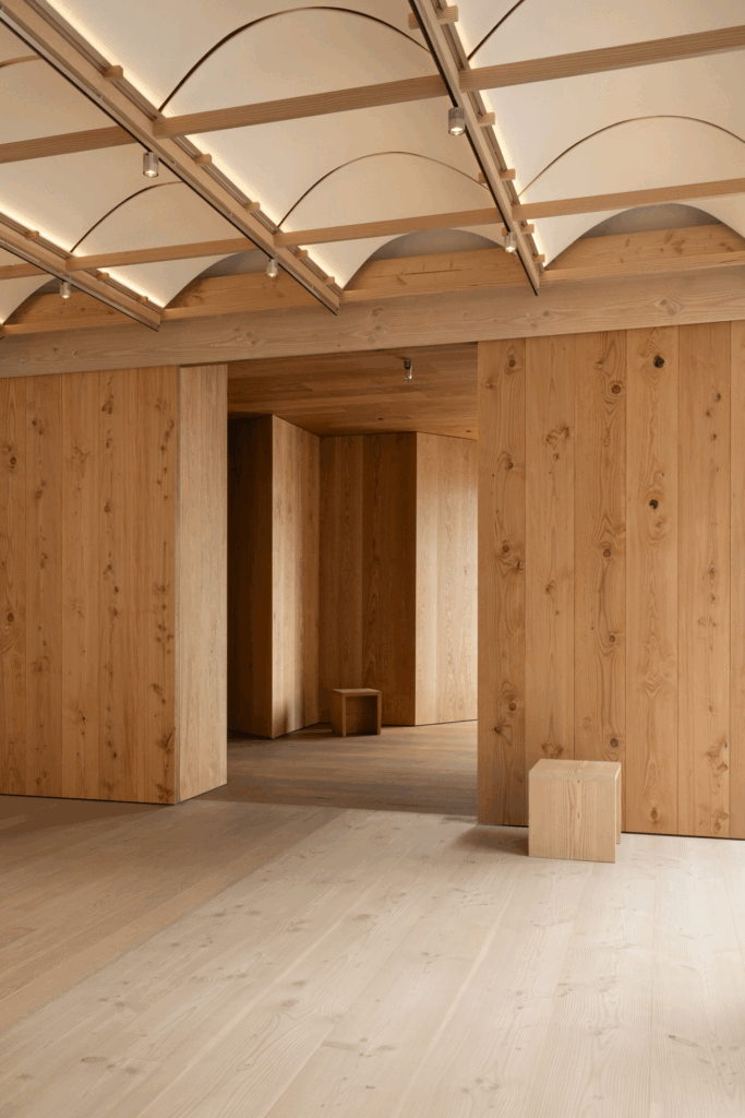 Minimalist interior with light wood floors, walls, and curved ceiling panels debuts at Milan Design Week 2026. Simple wooden blocks serve as seating, giving the open space a spacious and uncluttered feel.