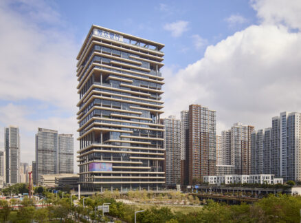 Modern high-rise building with staggered balconies stands among other tall buildings in an urban cityscape under a partly cloudy sky.