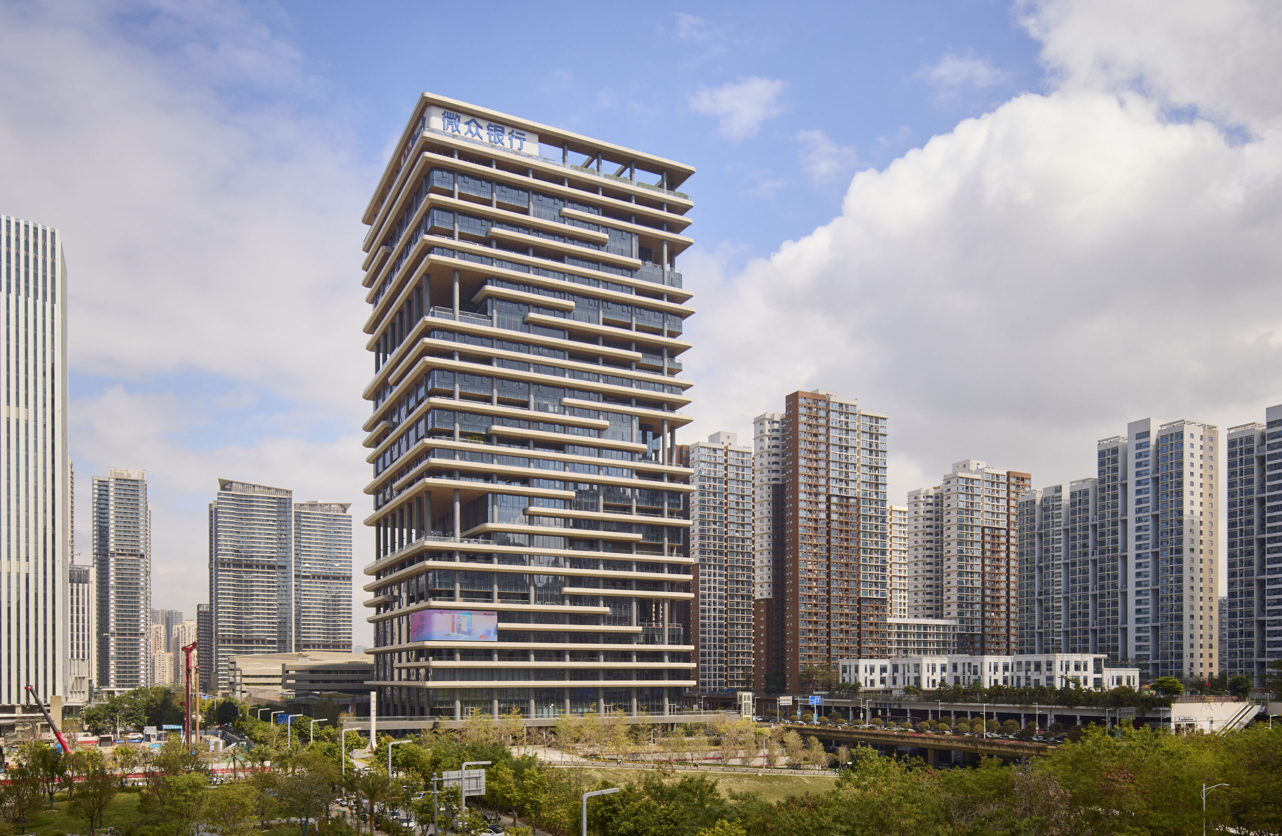 Modern high-rise building with staggered balconies stands among other tall buildings in an urban cityscape under a partly cloudy sky.