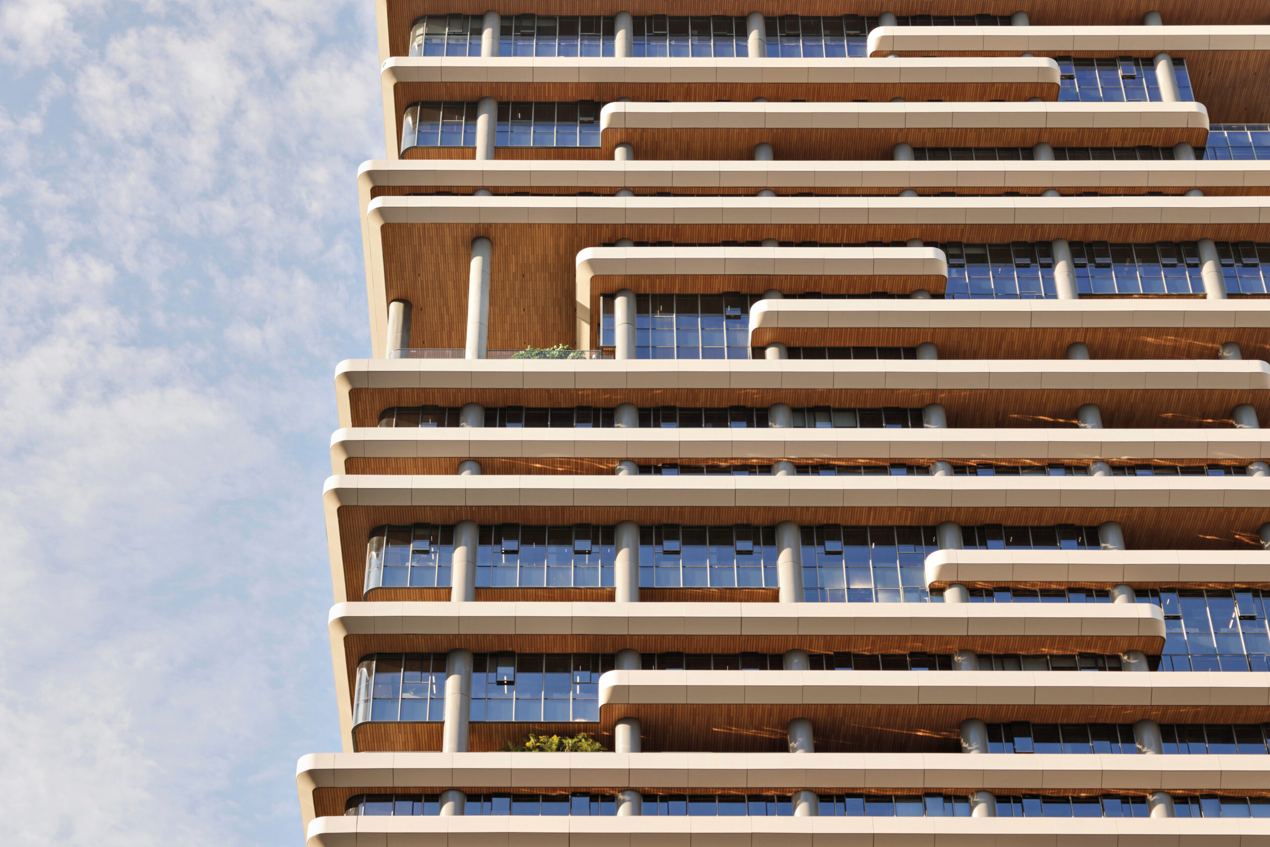 Modern building with staggered, layered balconies, wood accents, and large glass windows, viewed from below against a partly cloudy sky.