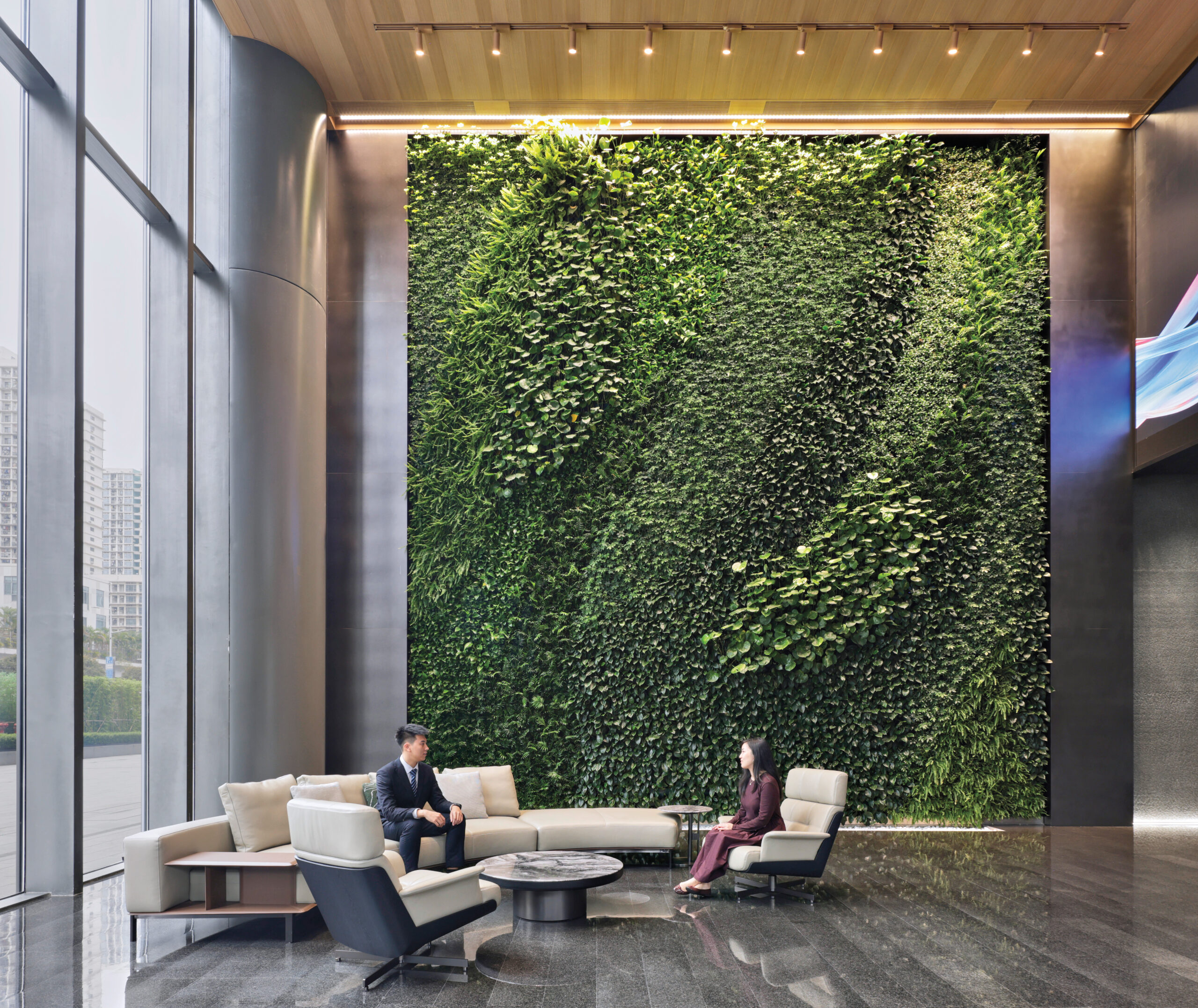 Two people sit on modern lounge chairs in a spacious lobby with a large green vertical garden wall and floor-to-ceiling windows.