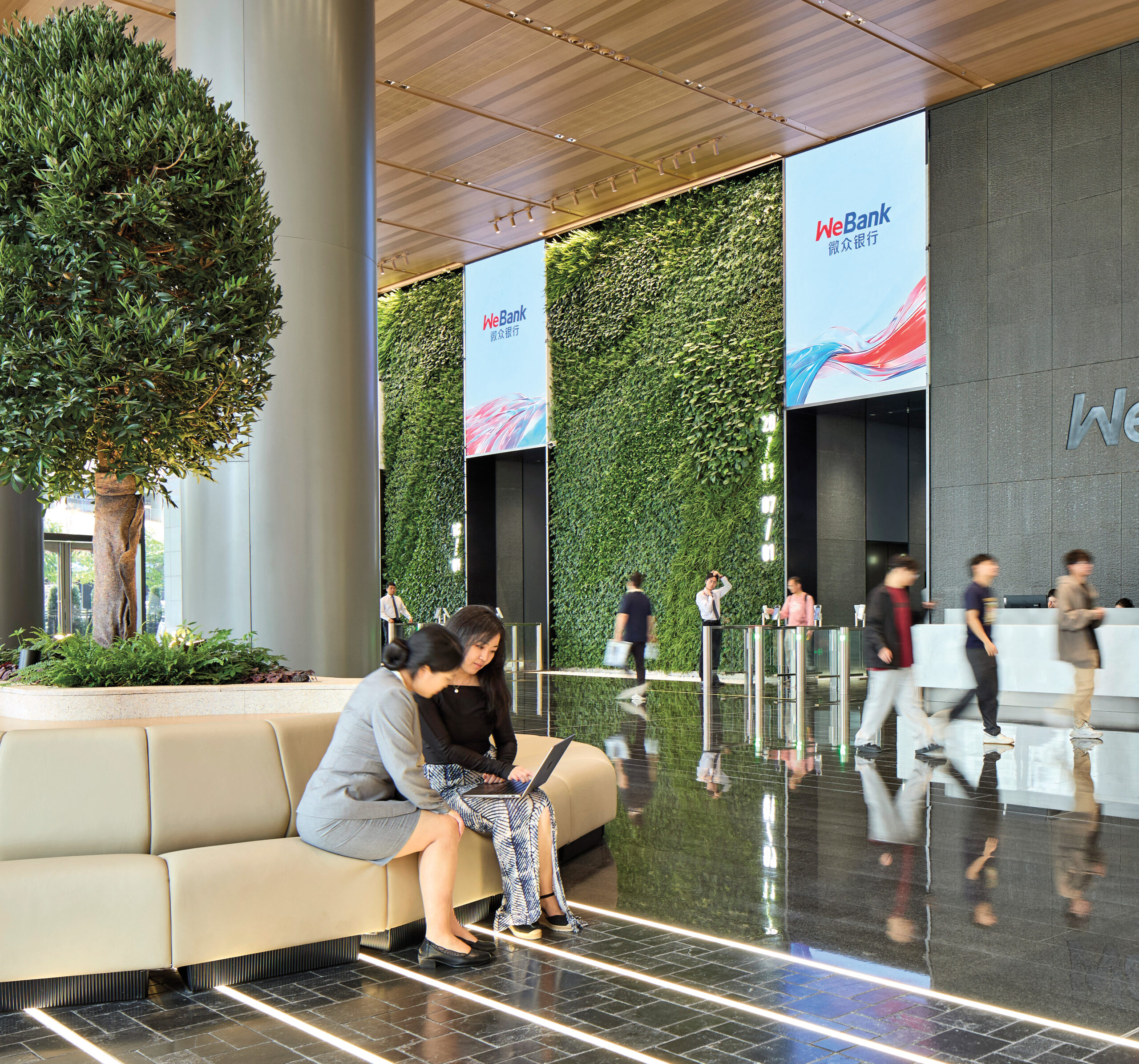 WeBank HQ: Modern bank lobby with a green wall, digital WeBank signs, people walking, and two women seated on a curved bench using a laptop.