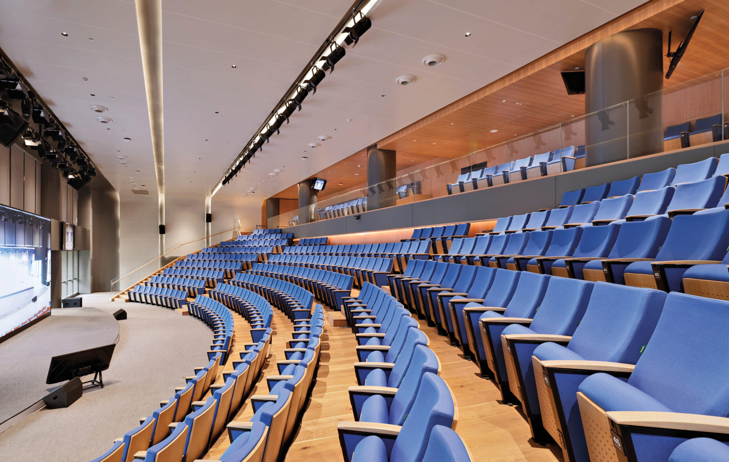 Modern auditorium with rows of blue seats, wooden accents, and a stage area at the front, all brightly lit with overhead lights.
