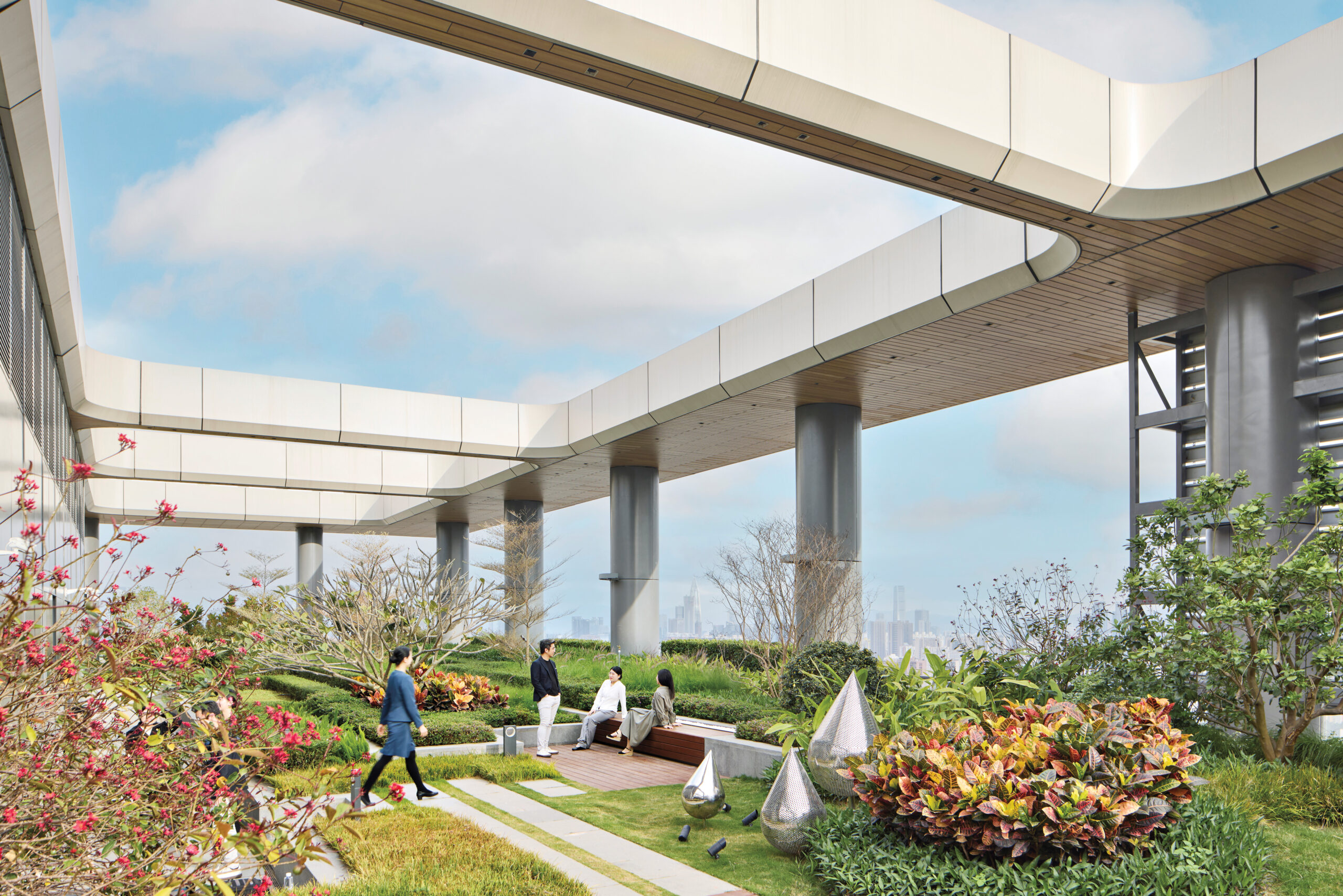 People walk and sit in a landscaped rooftop garden with large columns, modern architectural features, and city views in the background.
