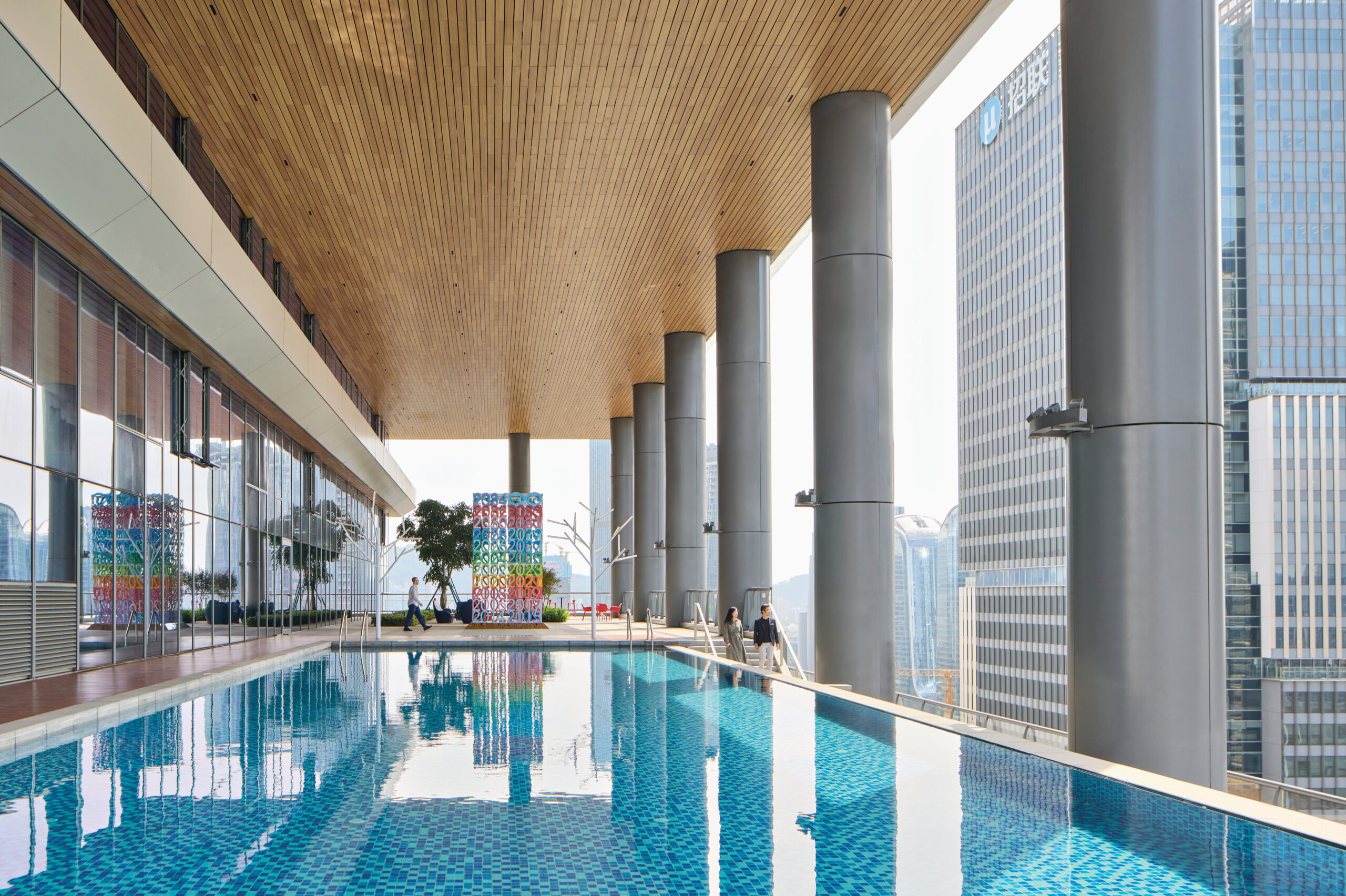 An elevated indoor pool with blue tiles, large structural columns, and a wooden ceiling, overlooking city skyscrapers through tall glass windows.