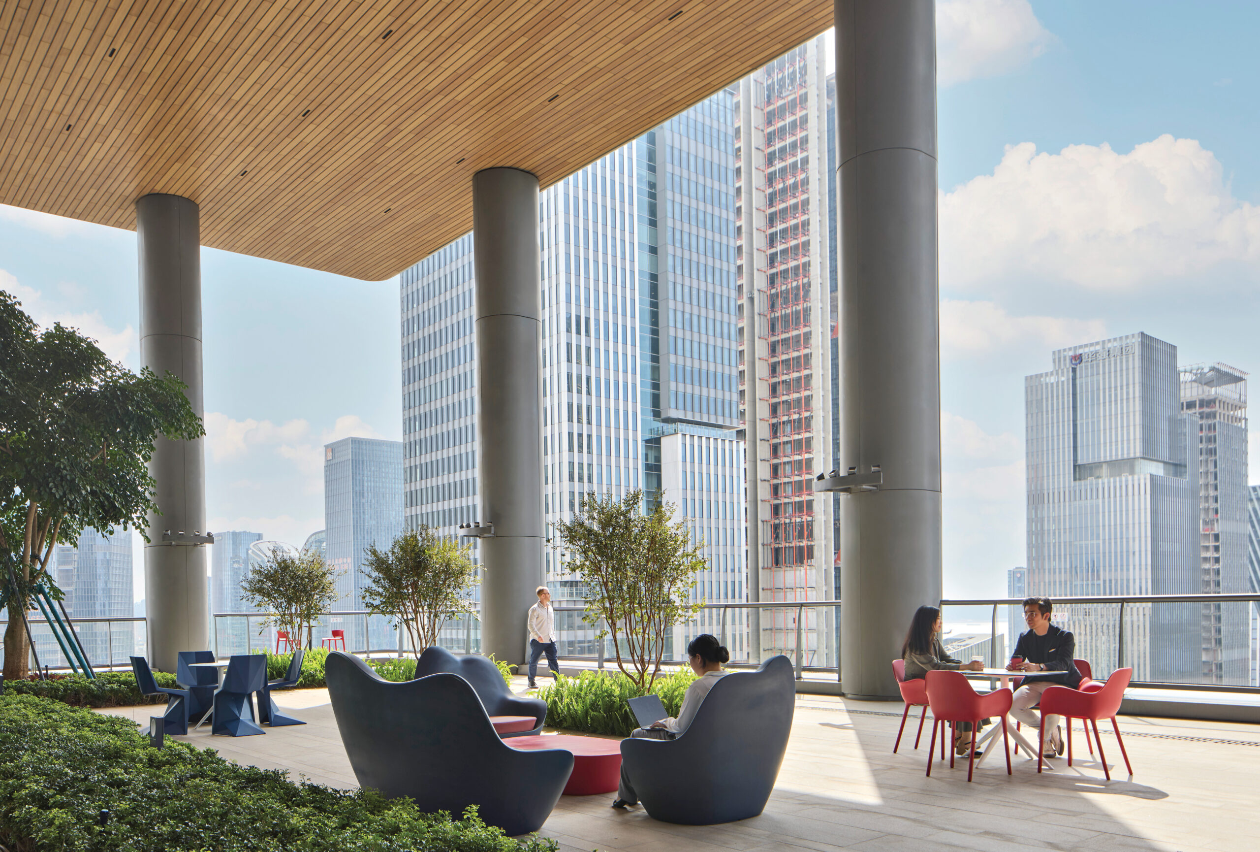 People sit and work or talk at tables and lounge chairs on a modern outdoor terrace with city skyscrapers in the background.