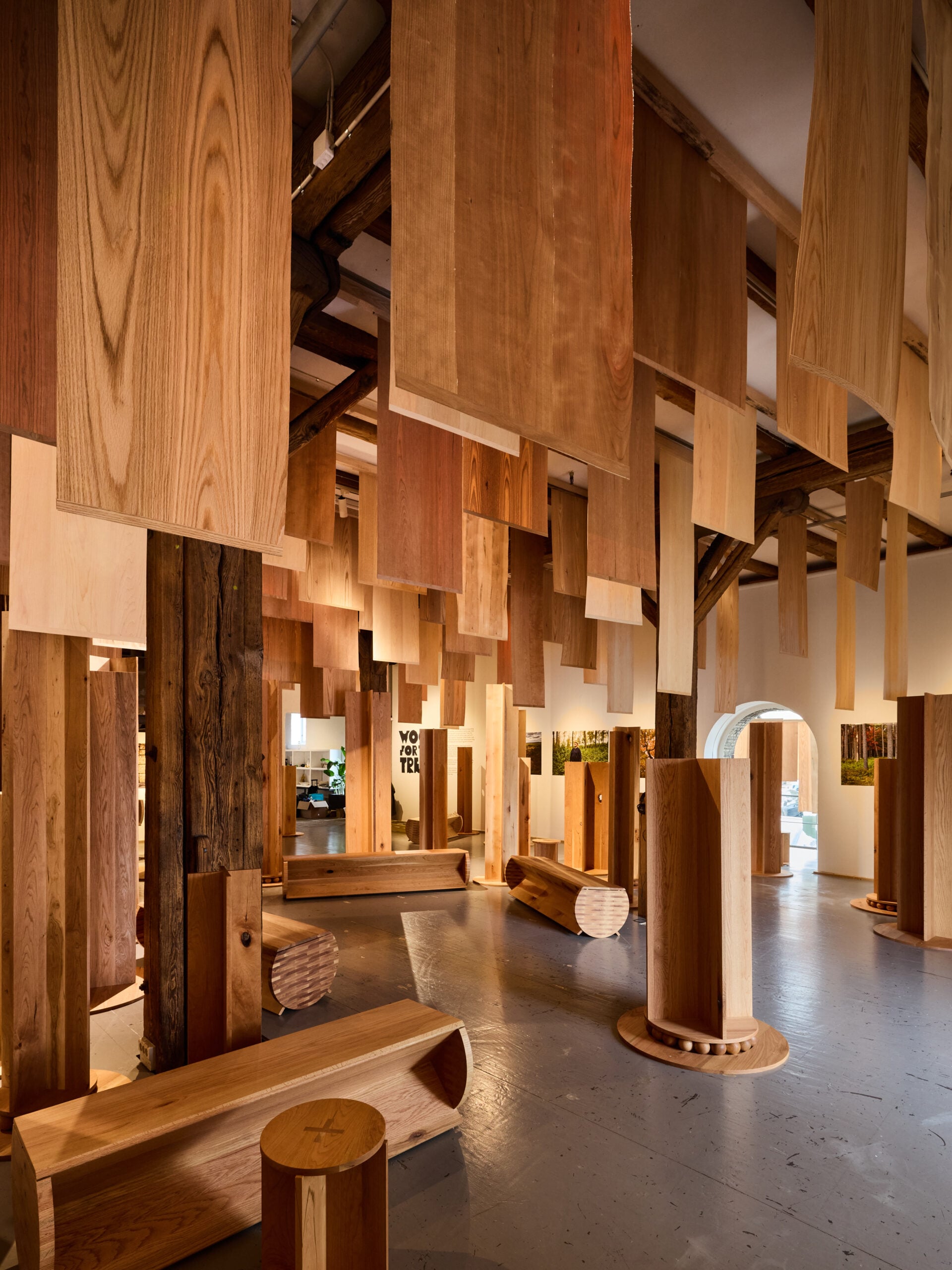 A room showcased at Milan Design Week 2026 features large vertical and horizontal wooden panels hanging from the ceiling, with wooden benches and pillars thoughtfully placed throughout the space.