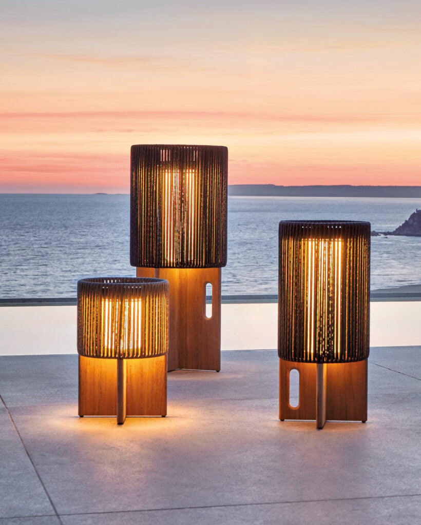 Three modern lantern-style outdoor lamps with wooden bases glow on a patio overlooking the ocean at sunset, creating a scene worthy of METROPOLISLikes.