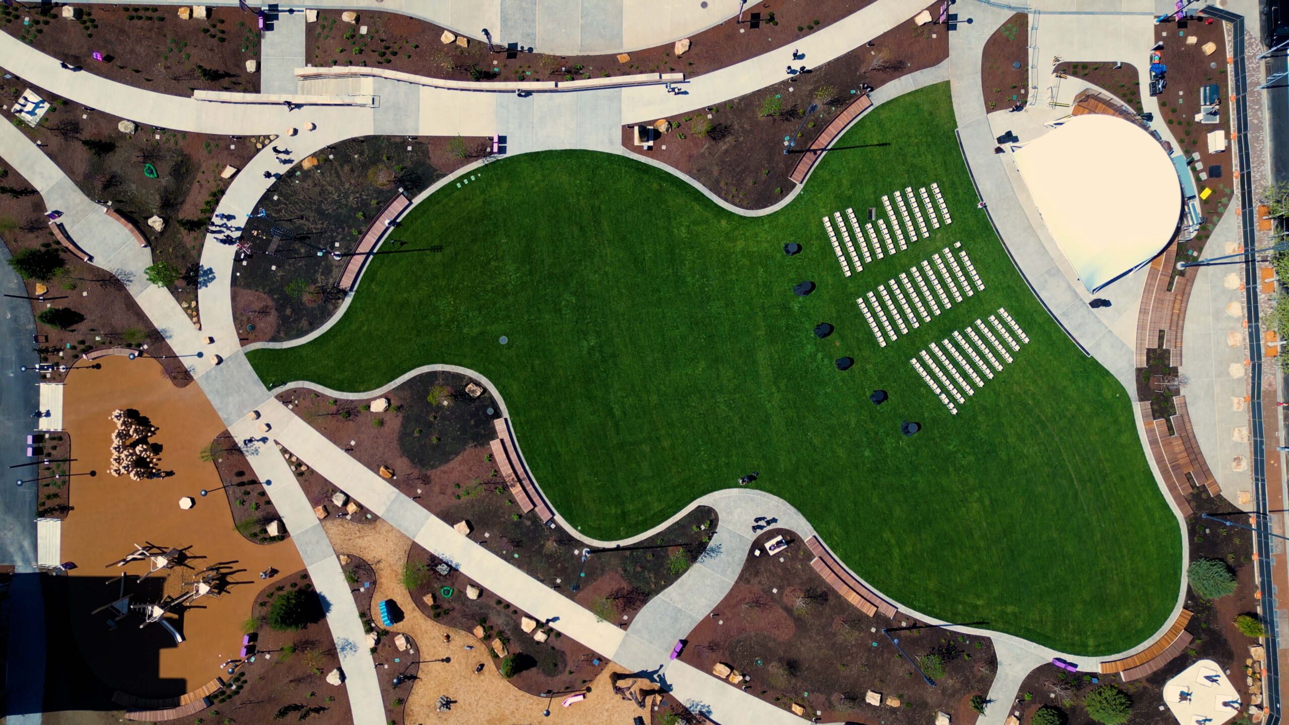 Aerial view of a park with a large green lawn, arranged rows of white chairs, winding paths, scattered people, and playground areas.