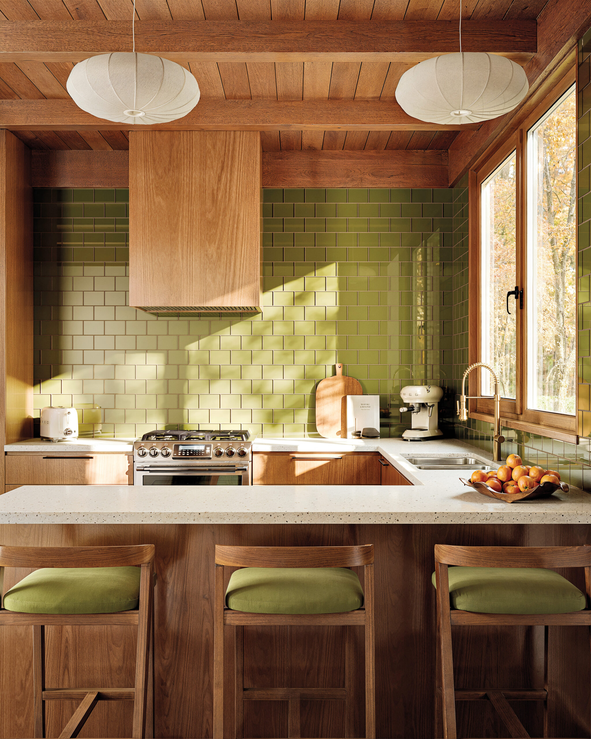 A modern kitchen with green tiled walls, wooden cabinets, a gas stove, pendant lights, and three bar stools with green cushions at a white countertop.