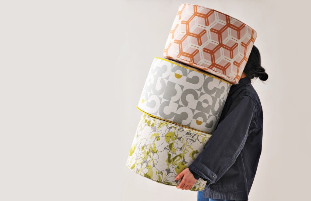 A person carries three large patterned cylinders stacked on top of each other, partially obscuring their face, against a plain background—an image sure to earn METROPOLISLikes.