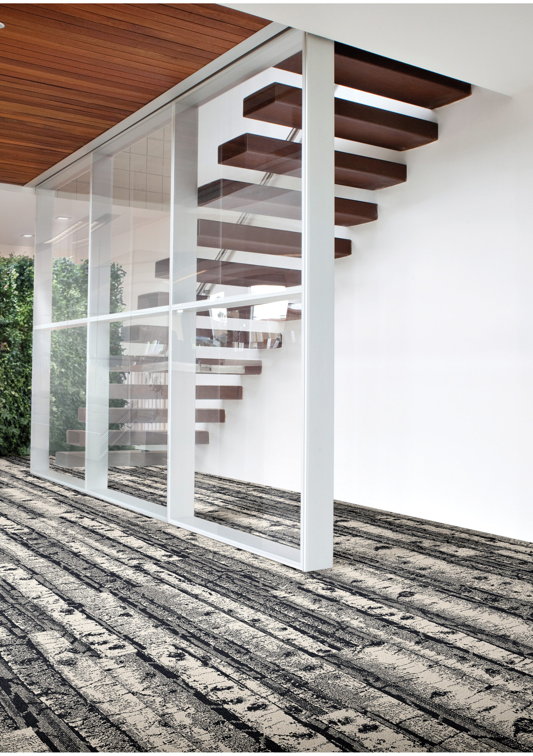 Open modern interior featuring floating wooden stairs behind a glass wall, with patterned gray and black carpeting and greenery in the background.