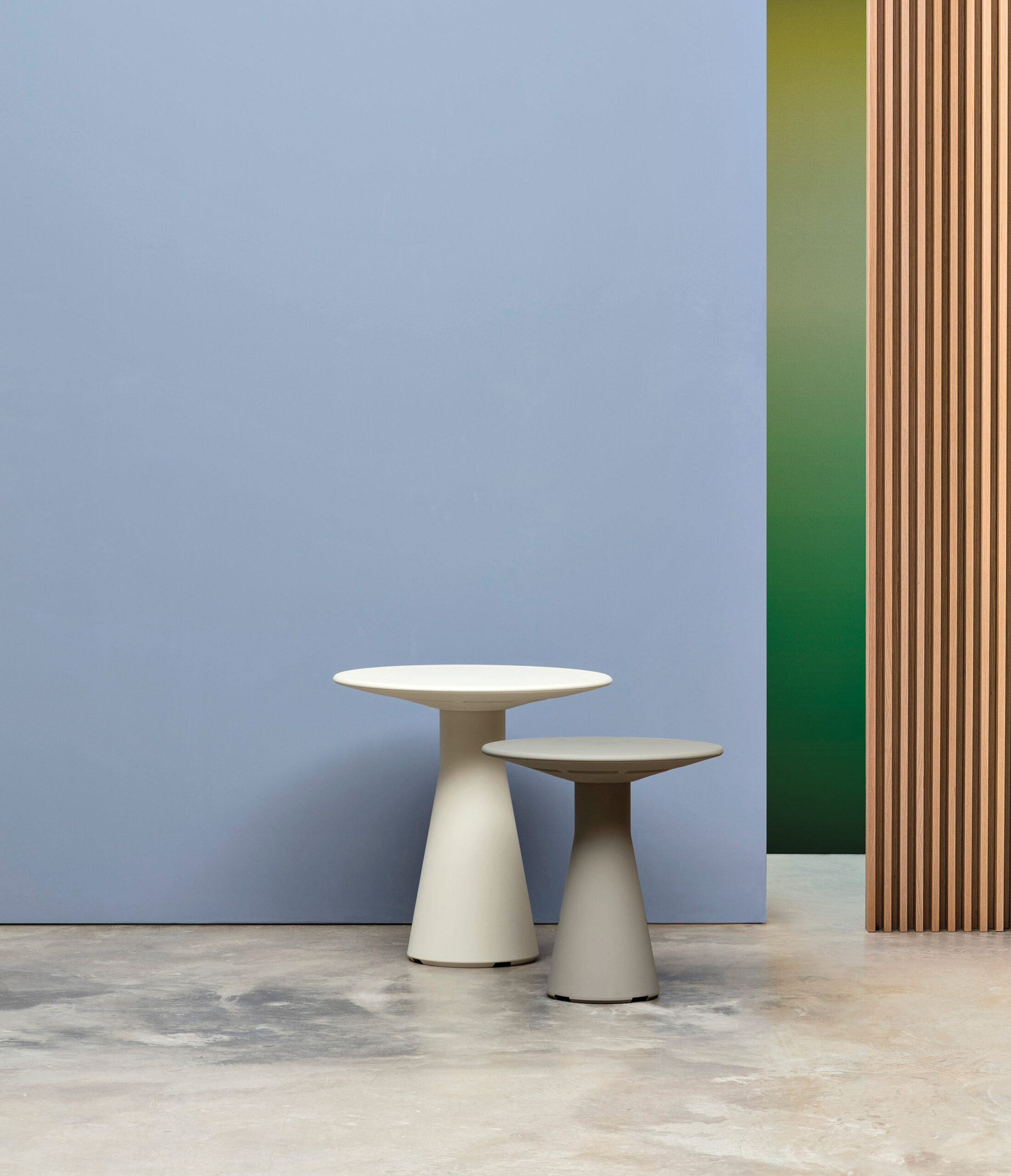 Two modern, minimalist round tables of different heights stand on a concrete floor against a blue wall, with a vertical wooden slat partition and a green gradient wall in the background.