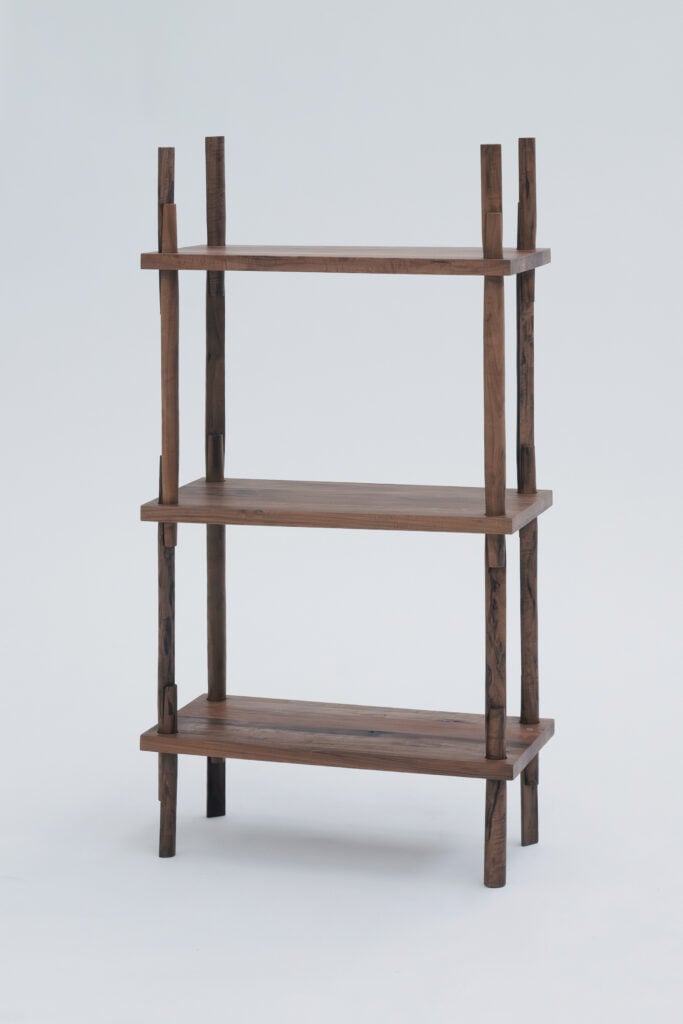 Three-tier wooden shelf with four vertical supports and a minimalist design, inspired by trends from Milan Design Week 2026, set against a plain light background.