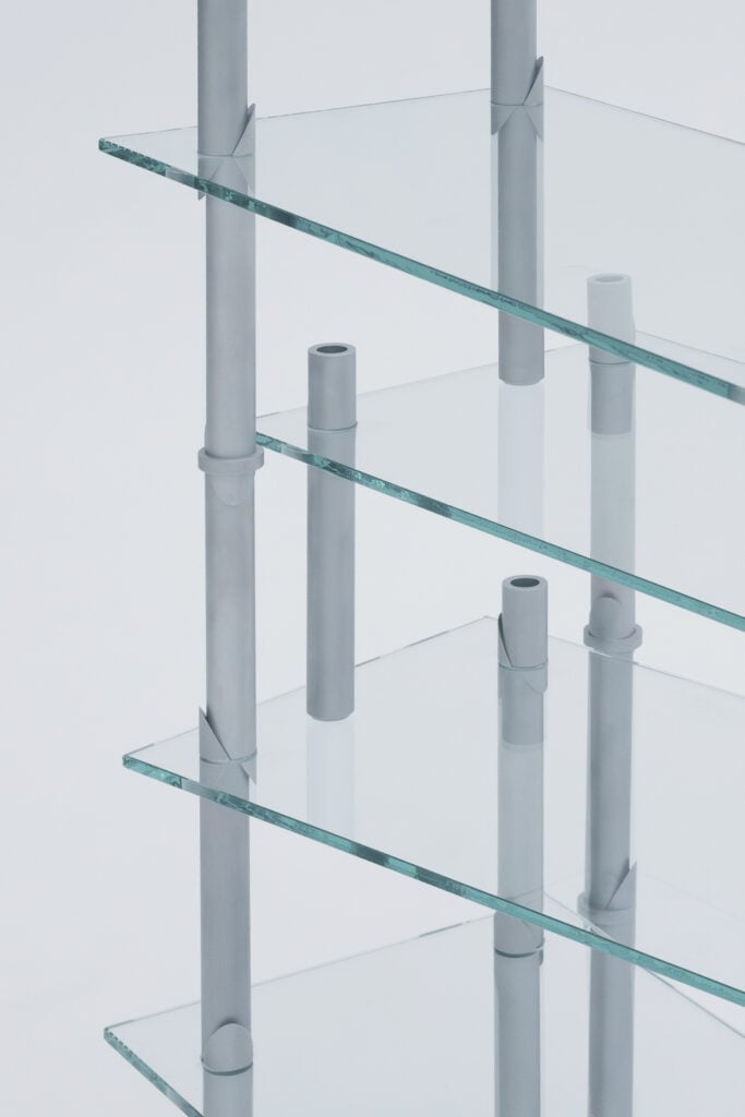 Close-up view of a modern shelving unit with transparent glass shelves and vertical metal supports, inspired by trends from Milan Design Week 2026, set against a plain background.