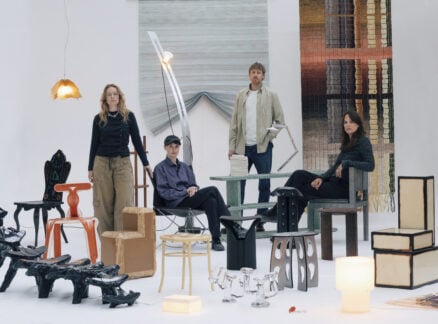 Four people pose among eclectic chairs, tables, and lamps in a minimalist studio with textured rugs as backdrops, capturing the creative spirit of Milan Design Week 2026.