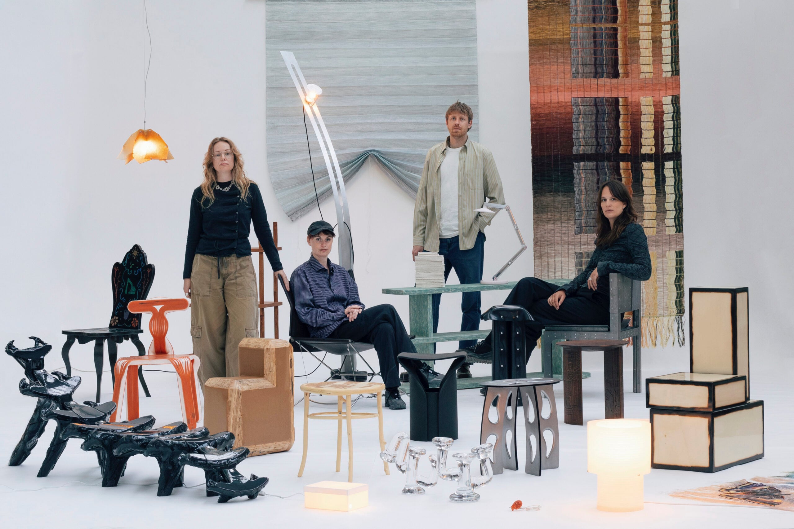 Four people pose among eclectic chairs, tables, and lamps in a minimalist studio with textured rugs as backdrops, capturing the creative spirit of Milan Design Week 2026.