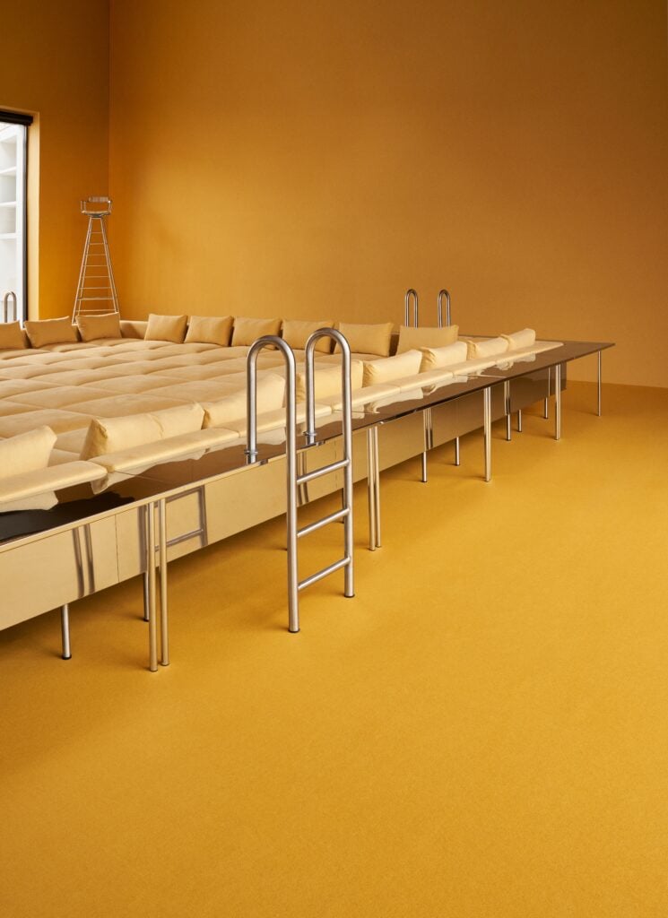 A room with a large, raised platform featuring cushioned yellow mats, metallic ladders, and a monochromatic yellow color scheme, inspired by the creative spirit of Milan Design Week 2026.