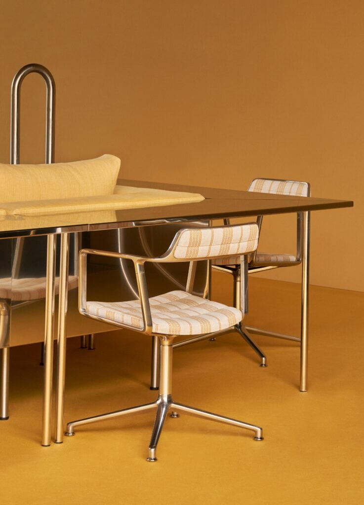 Modern dining area with a reflective metal table, two metal-framed chairs with striped cushions, and a yellow pillow, set against a yellow background—perfect inspiration for Milan Design Week 2026.