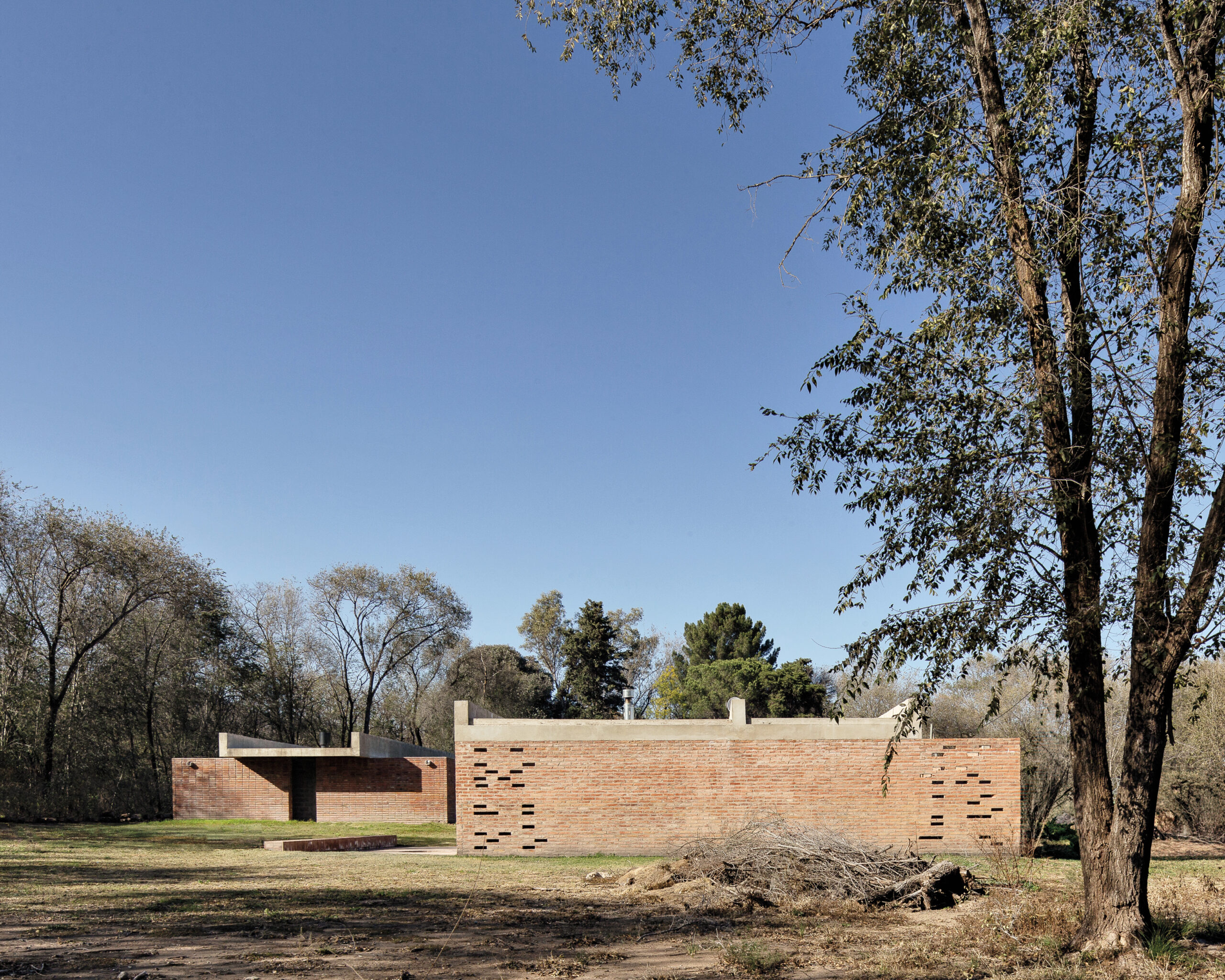 A modern, single-story brick building by Neri&Hu with rectangular cutouts stands in a grassy, wooded area under a clear blue sky.