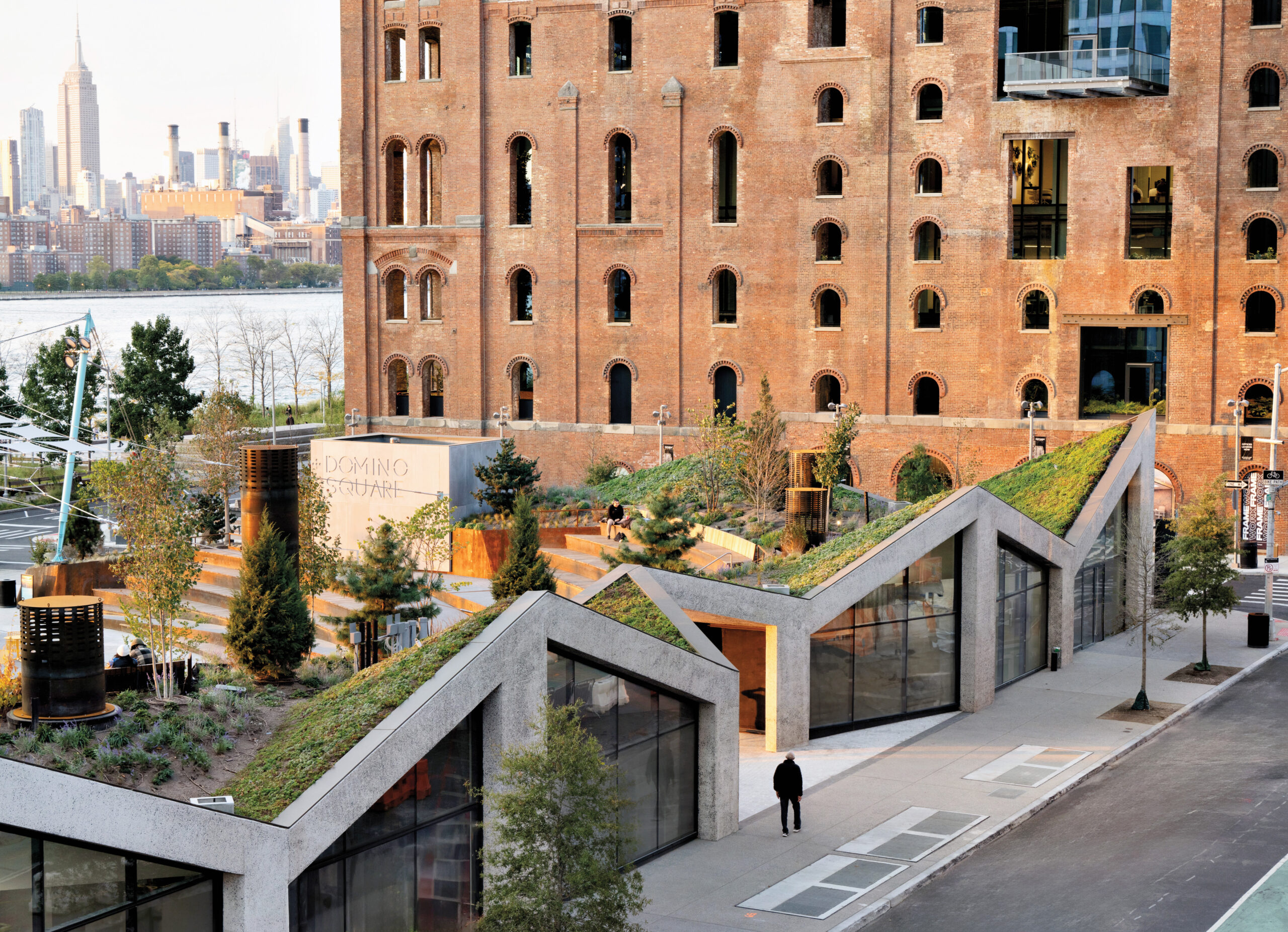 A modern Neri&Hu building with green roofs and glass walls sits in front of a historic brick structure, with a city skyline and river visible in the background.