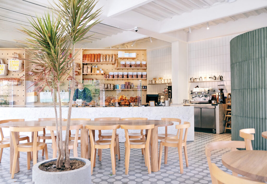 Bright, modern café interior with wooden tables and chairs, potted plant, open shelving, Latin Matter coffee equipment, and a person working behind the counter.