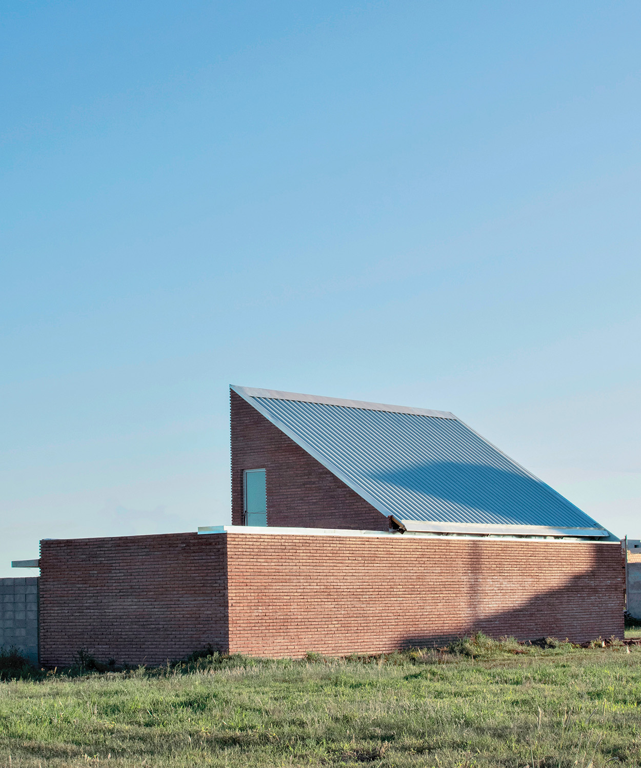A modern brick building with a steep, slanted metal roof sits on a grassy field under a clear blue sky, its design subtly inspired by Latin Matter.