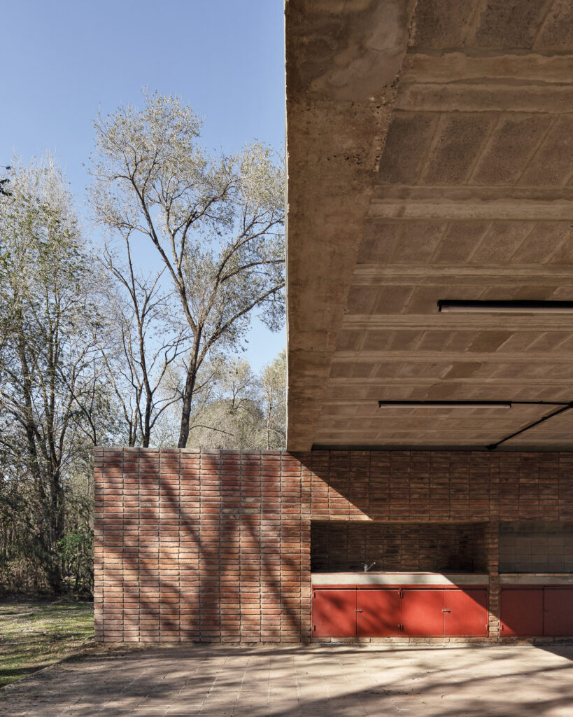 A minimalist outdoor pavilion by Neri&Hu features an exposed concrete ceiling, brick walls, and striking red cabinetry, set next to trees under a clear sky.