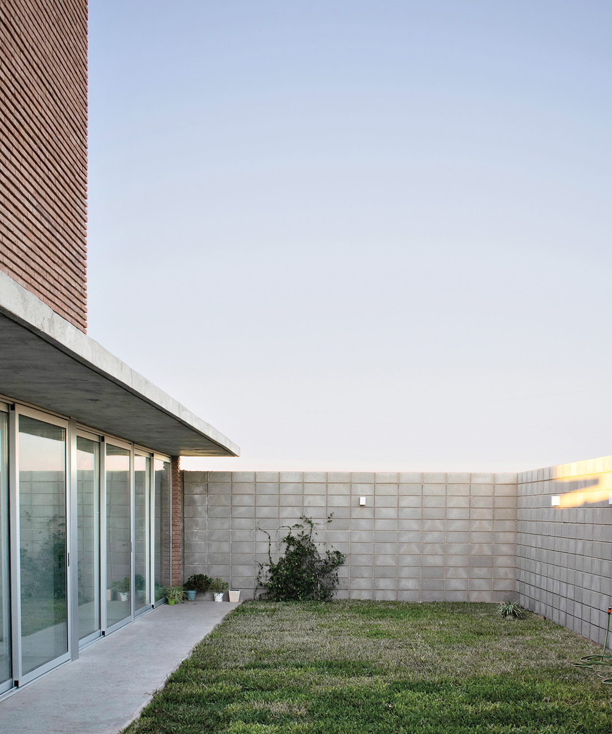 Modern outdoor patio with a glass sliding door, concrete overhang, cinder block walls, potted plants, and a grassy lawn under a clear sky—an inviting retreat inspired by Latin Matter design.
