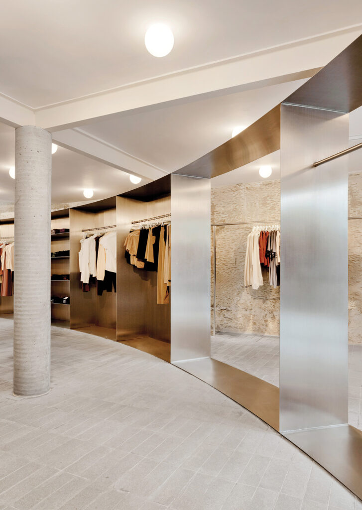 Minimalist clothing store interior by Latin Matter, featuring stainless steel racks, neutral-toned garments, a concrete column, textured wall, and bright ceiling lights.