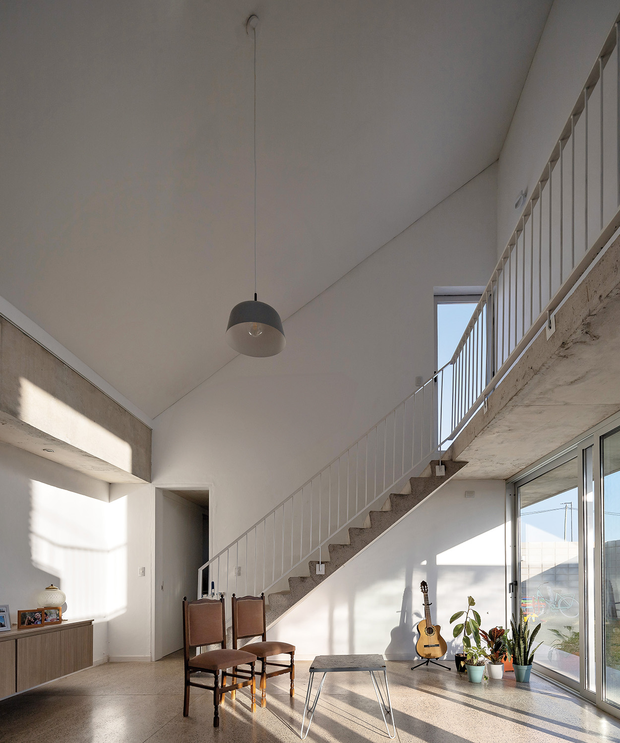 Modern living room with high ceiling, minimalist furniture, a staircase leading to an upper floor, large windows, potted plants, and a guitar by the railing—an inviting space inspired by Latin Matter design.