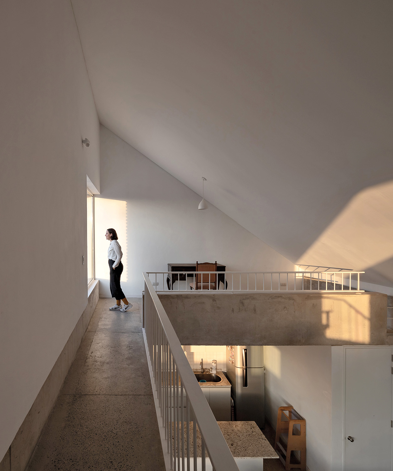 A person stands by a large window in a minimalist Latin Matter interior with slanted ceilings, white walls, and a concrete walkway overlooking a kitchen and dining area below.