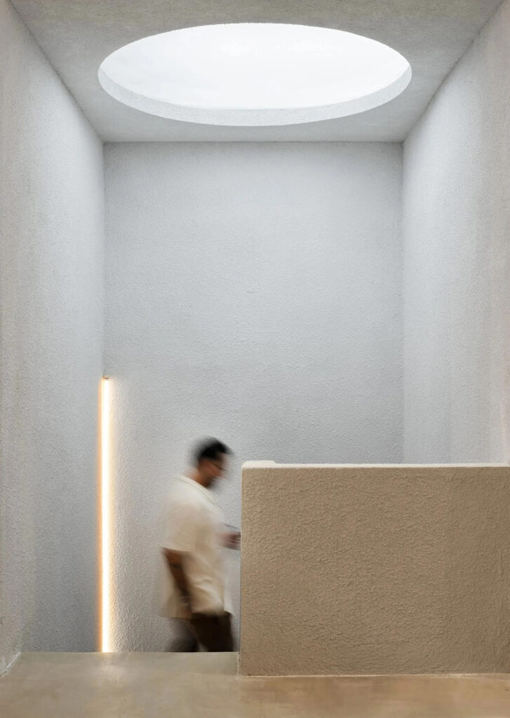 A person walks past a textured, light-colored wall in a minimalistic room with a circular skylight, a vertical strip light, and subtle hints of Latin Matter in the architectural details.