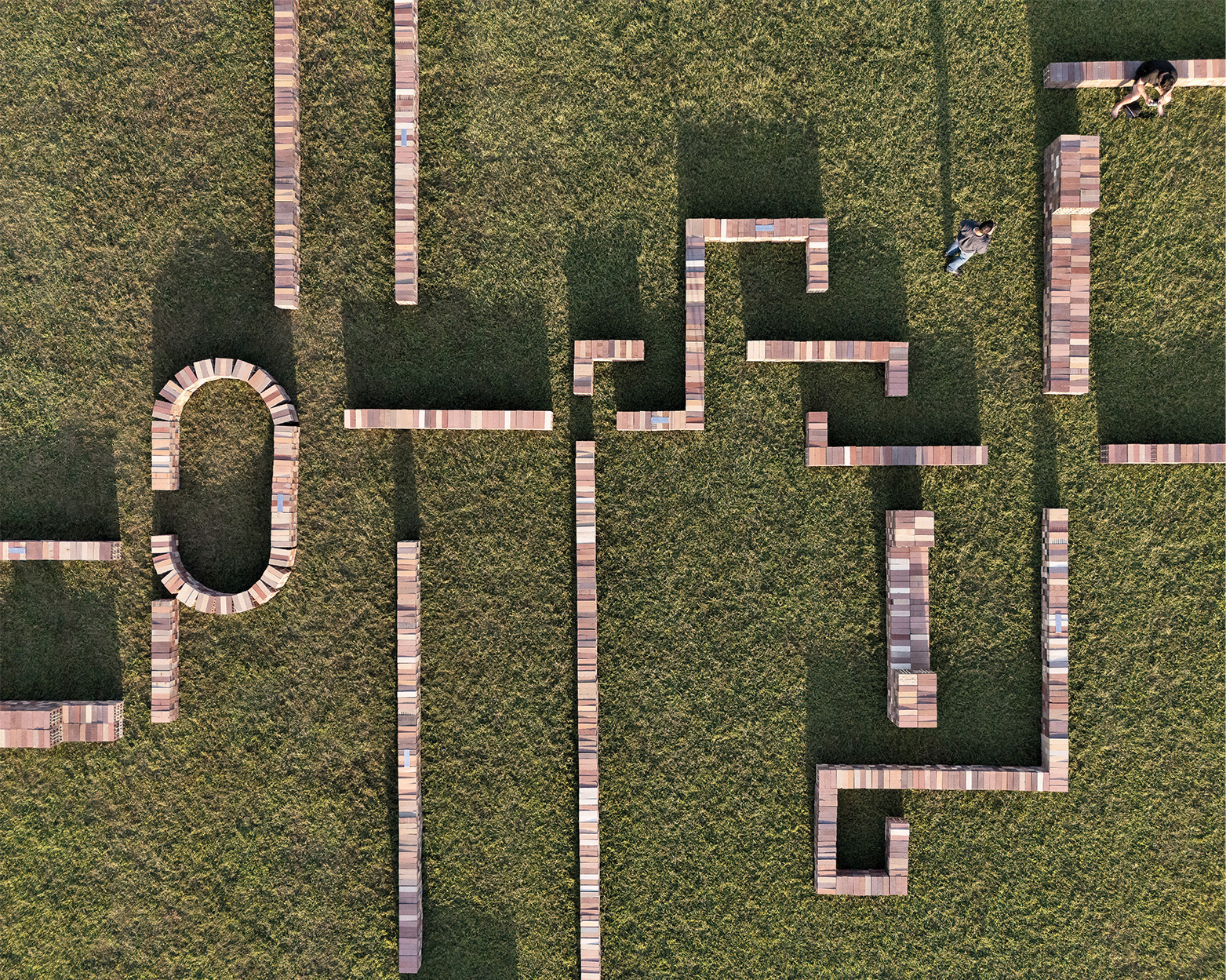 Aerial view of a grassy field with wooden planks arranged in geometric patterns; two people, engaged in a Latin Matter project, stand among the planks casting long shadows.